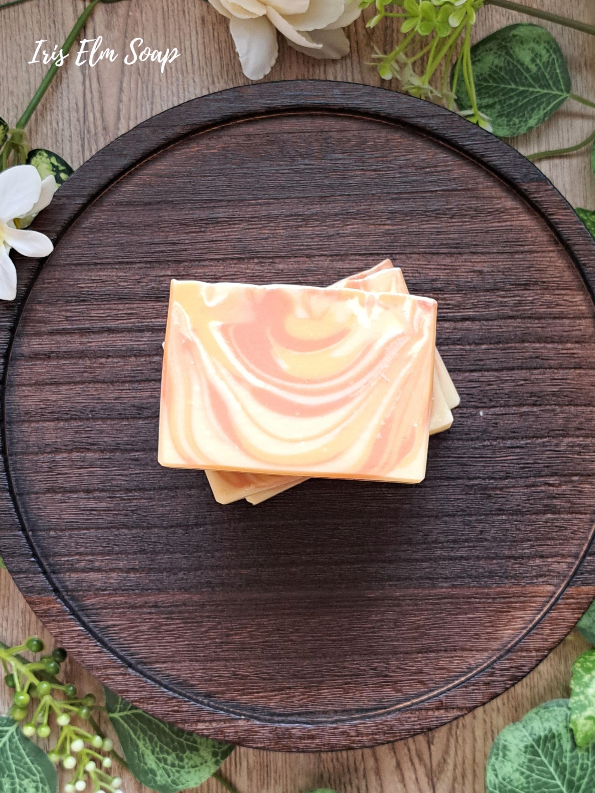 Stack of mango tangerine handmade soap bars with marbled design on a wooden board, surrounded by greenery.