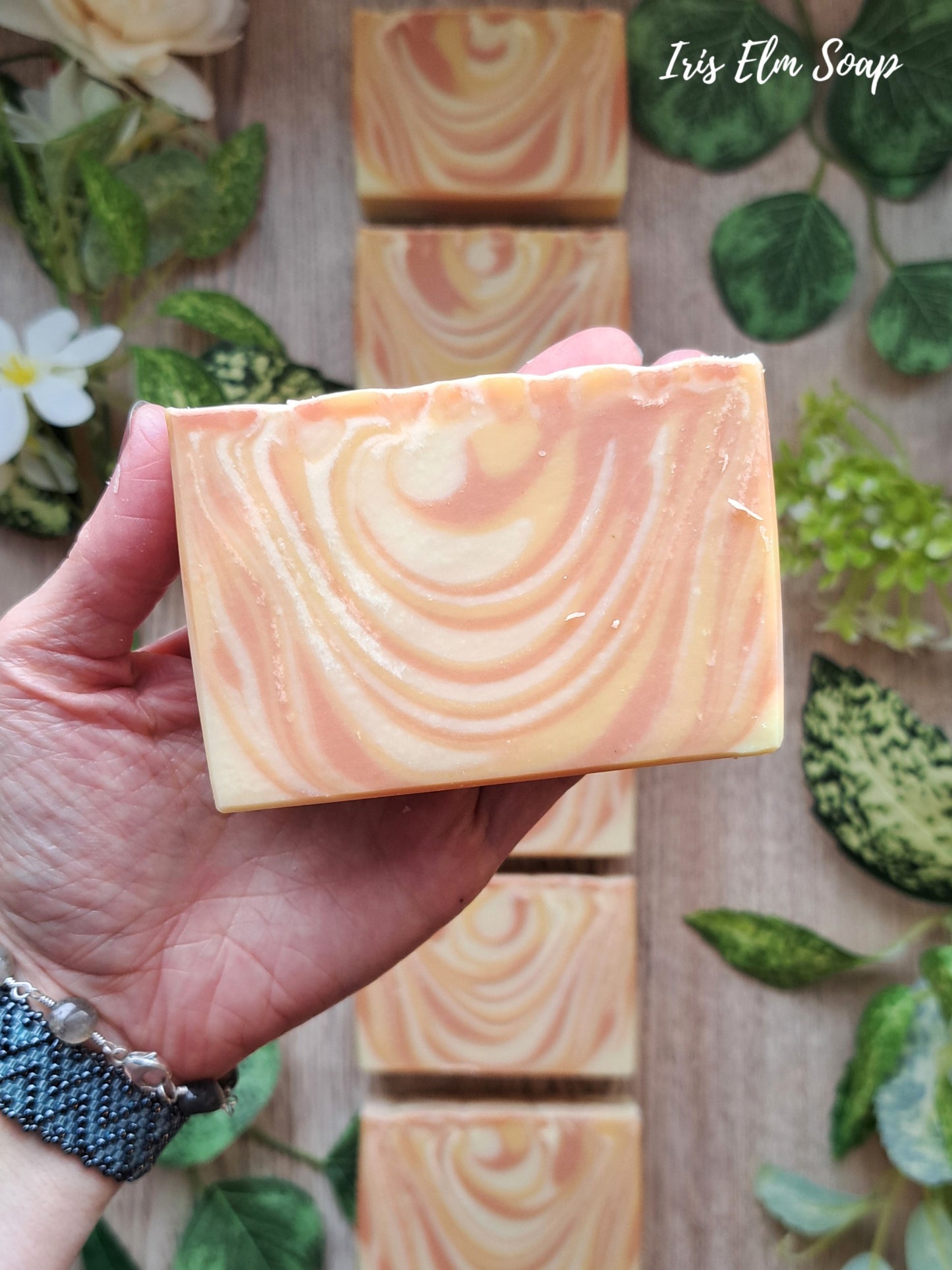 Hand holding a bar of mango tangerine handmade soap with a swirled pattern, set against a wooden background with more soap bars and greenery.