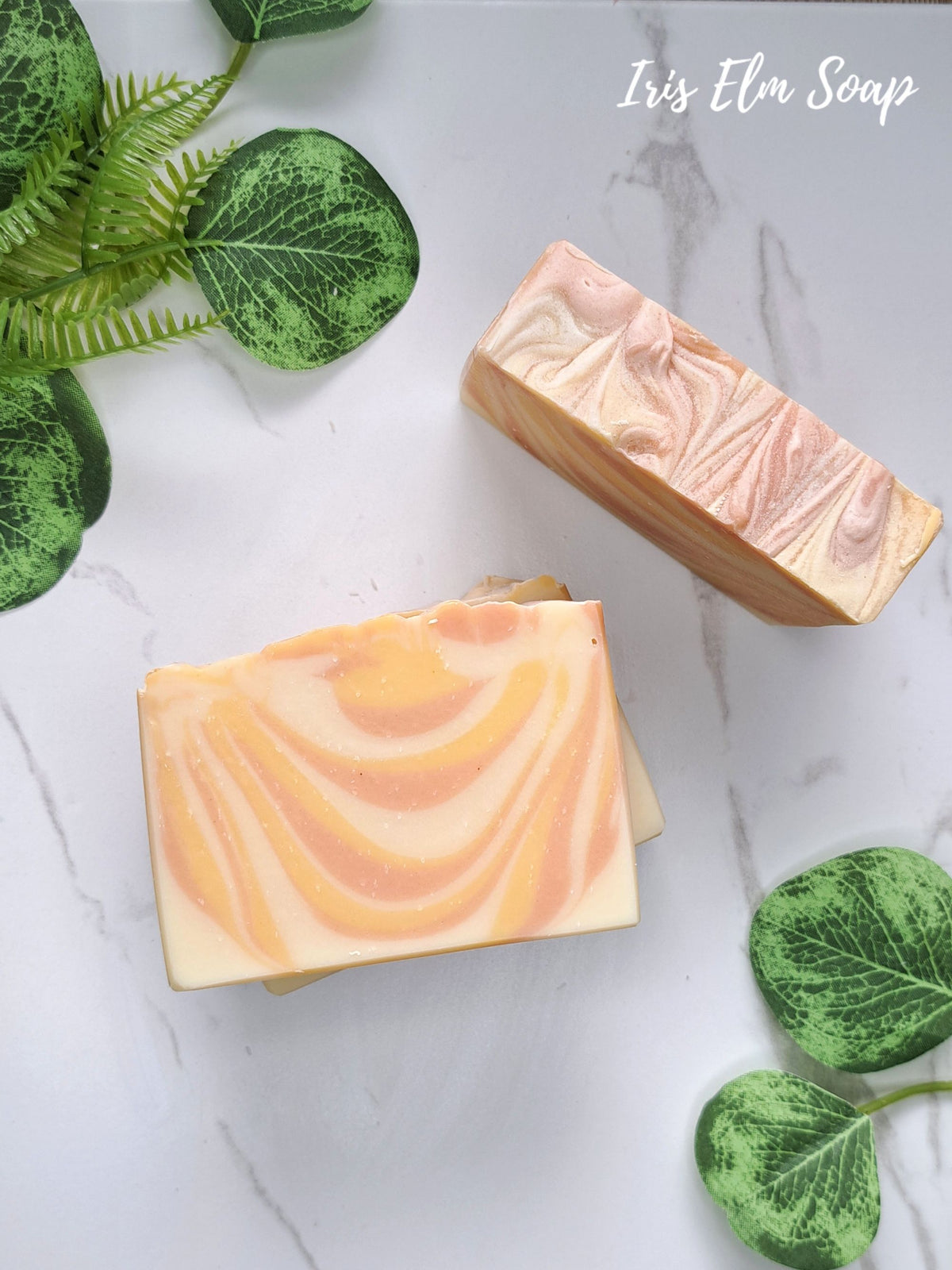 Two rectangular bars of swirled mango tangerine handmade soap with peach, orange, and cream tones arranged on a white marble surface with green decorative leaves. The text 'Iris Elm Soap' appears in the top right.