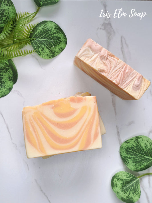 Two rectangular bars of swirled mango tangerine handmade soap with peach, orange, and cream tones arranged on a white marble surface with green decorative leaves. The text 'Iris Elm Soap' appears in the top right.