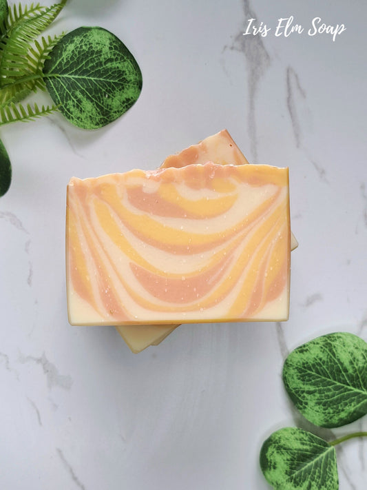 Two stacked bars of swirled yellow and orange mango tangerine handmade soap with fine glitter, styled on a white marble surface with green leaves. The text 'Iris Elm Soap' is visible in the top right corner.