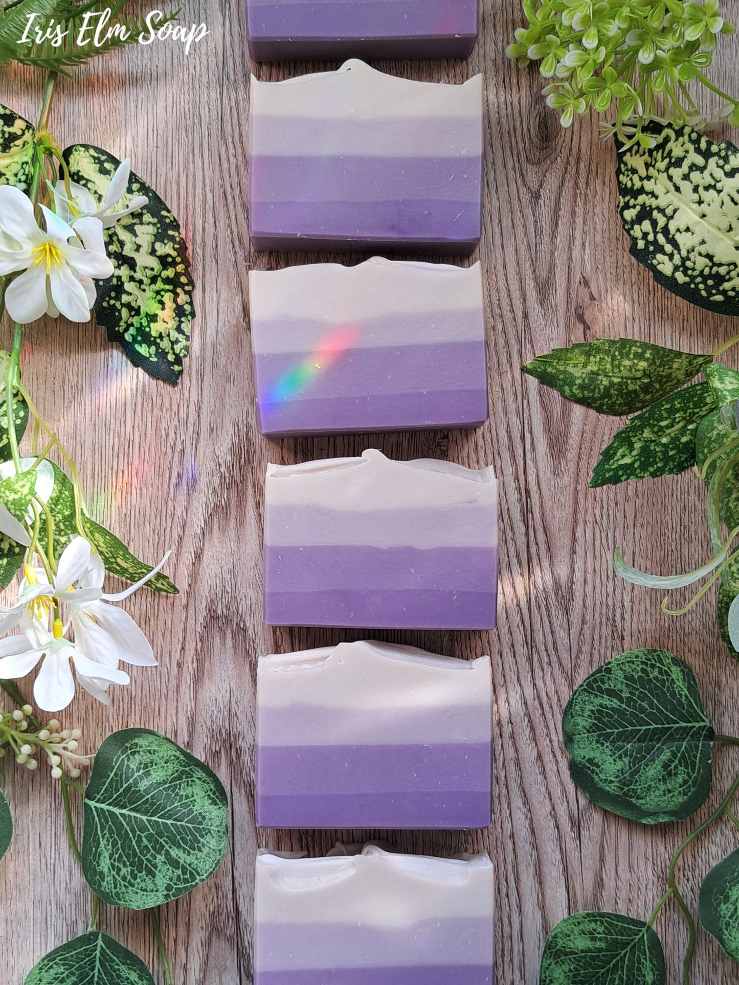 Gradient purple and white French lilac handmade floral soap bars on a wooden surface with green leaves and flowers.