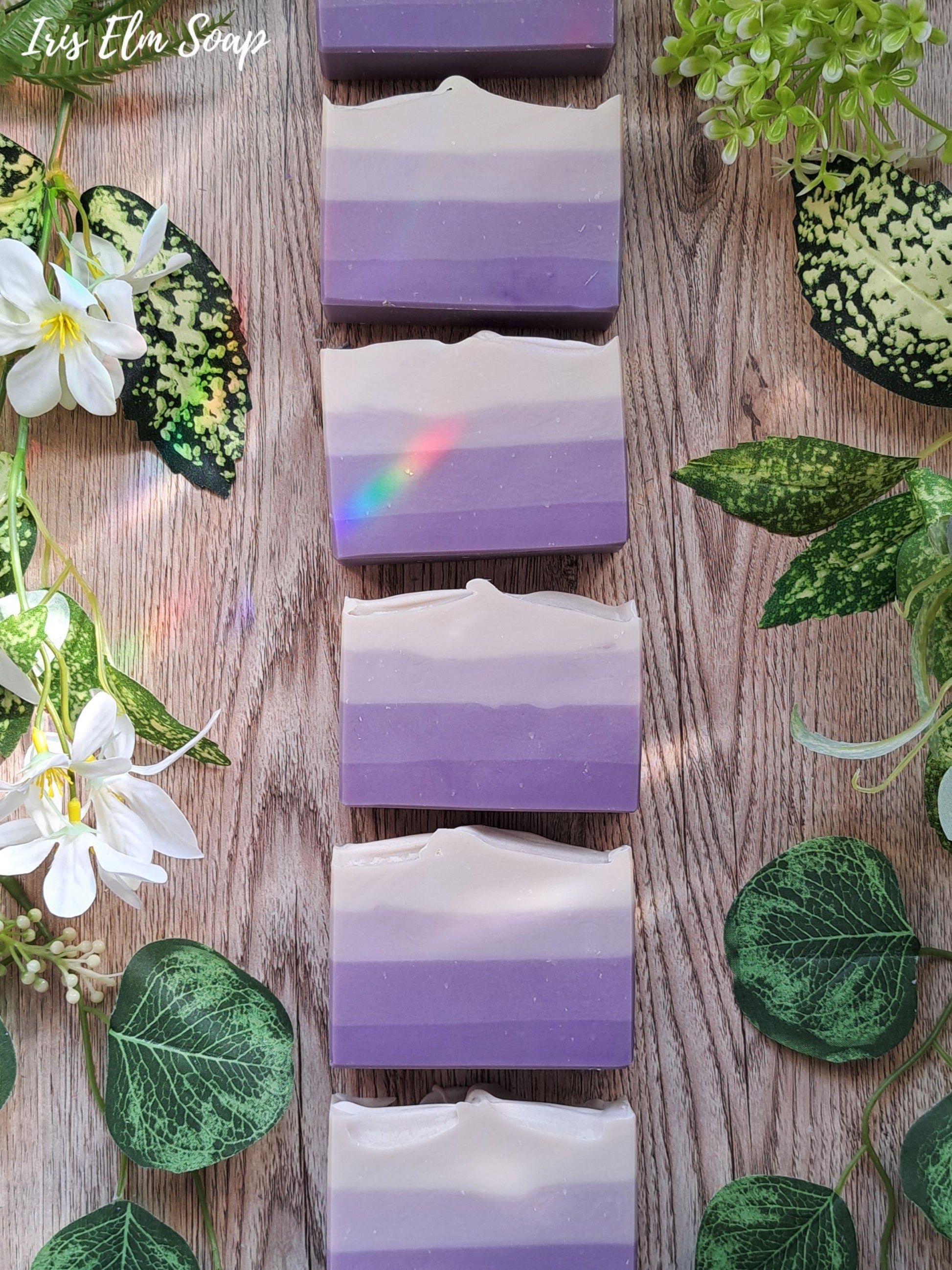 Gradient purple and white French lilac handmade floral soap bars on a wooden surface with green leaves and flowers.