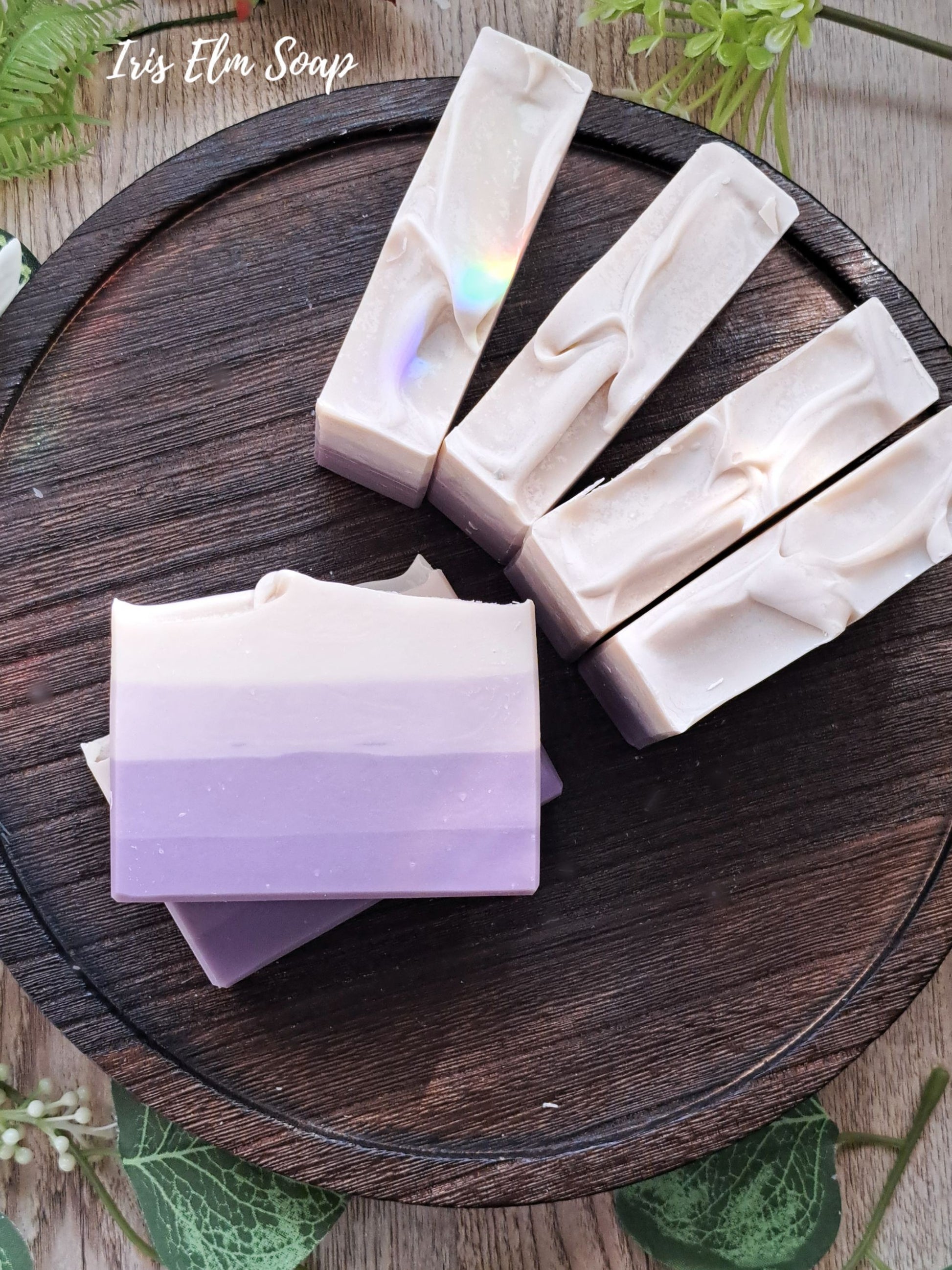 Three bars of French lilac handmade soap with different color layers on a wooden surface, with 'Iris Elm Soap' branding.