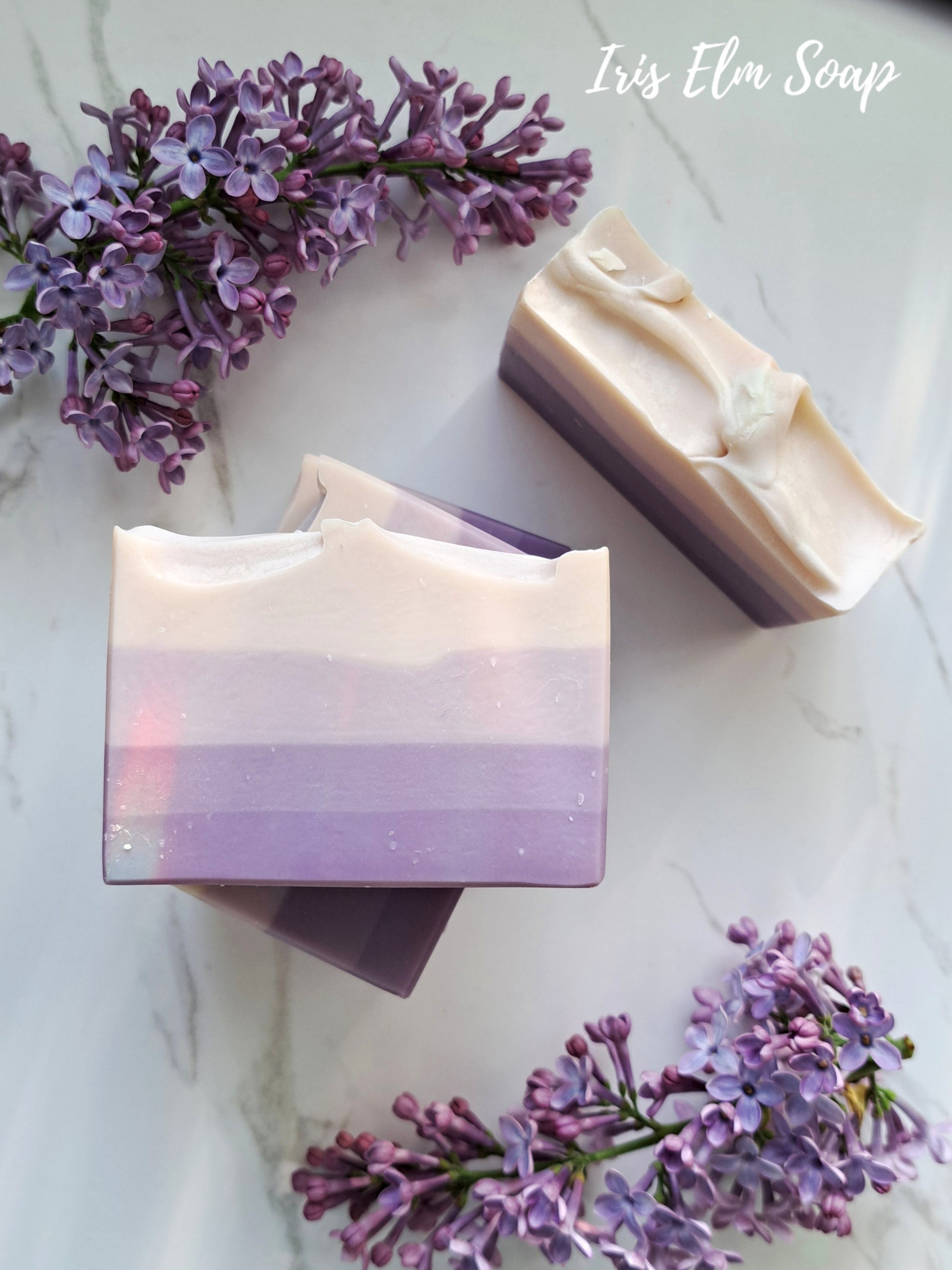 Two bars of French lilac handmade soap with a gradient design surrounded by purple flowers on a light background, with 'Iris Elm Soap' branding.