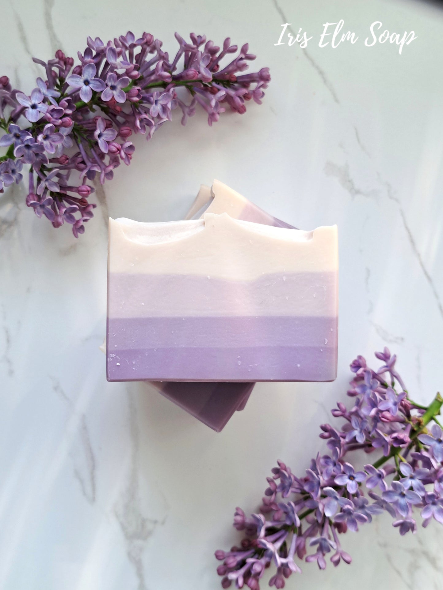 Bar of purple French lilac handmade soap with a gradient design surrounded by purple flowers on a light background