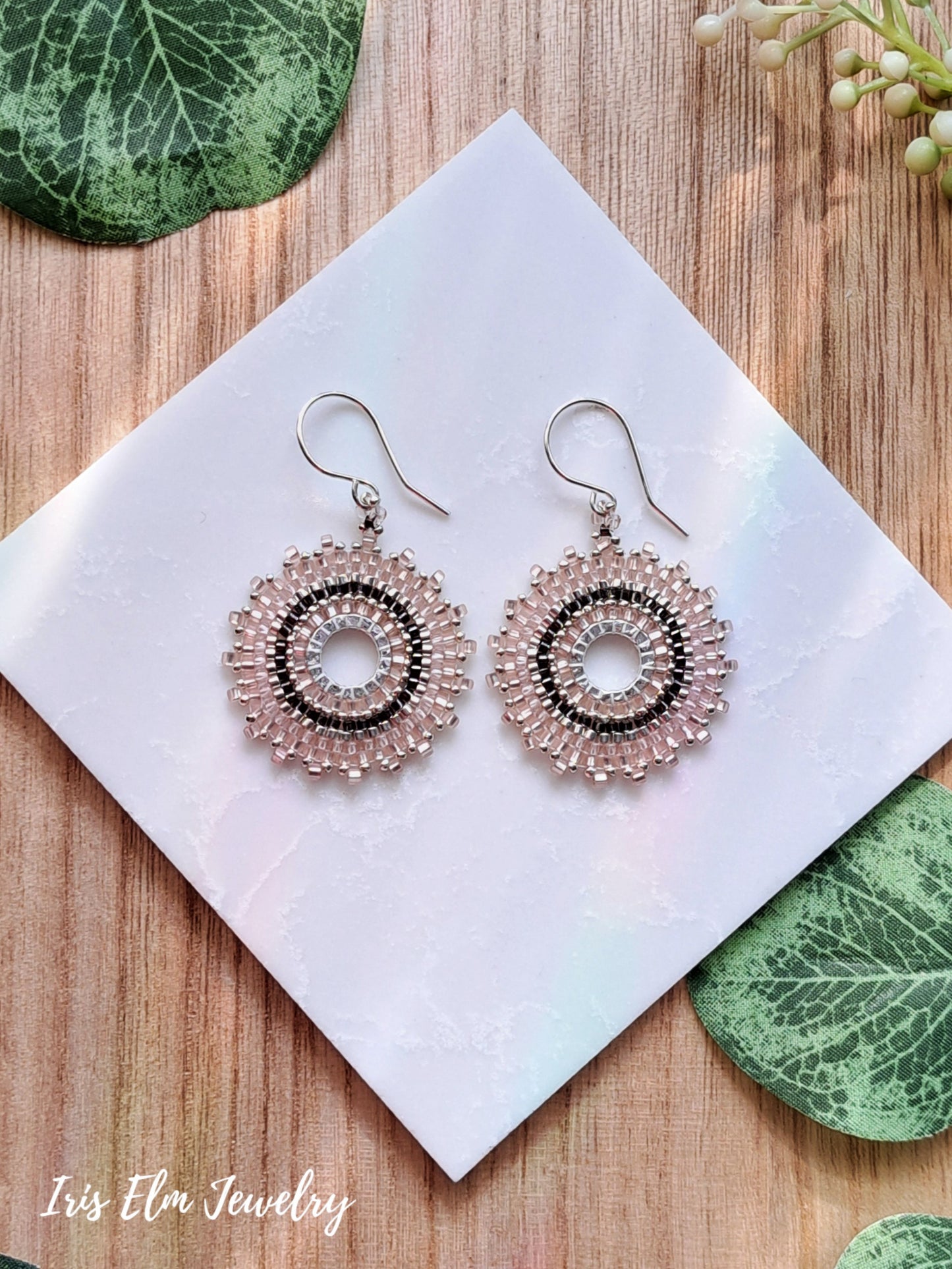 Pair of beaded pale pink seed bead hoop earrings on a white card with green leaves on a wooden surface, featuring 'Iris Elm Jewelry'.
