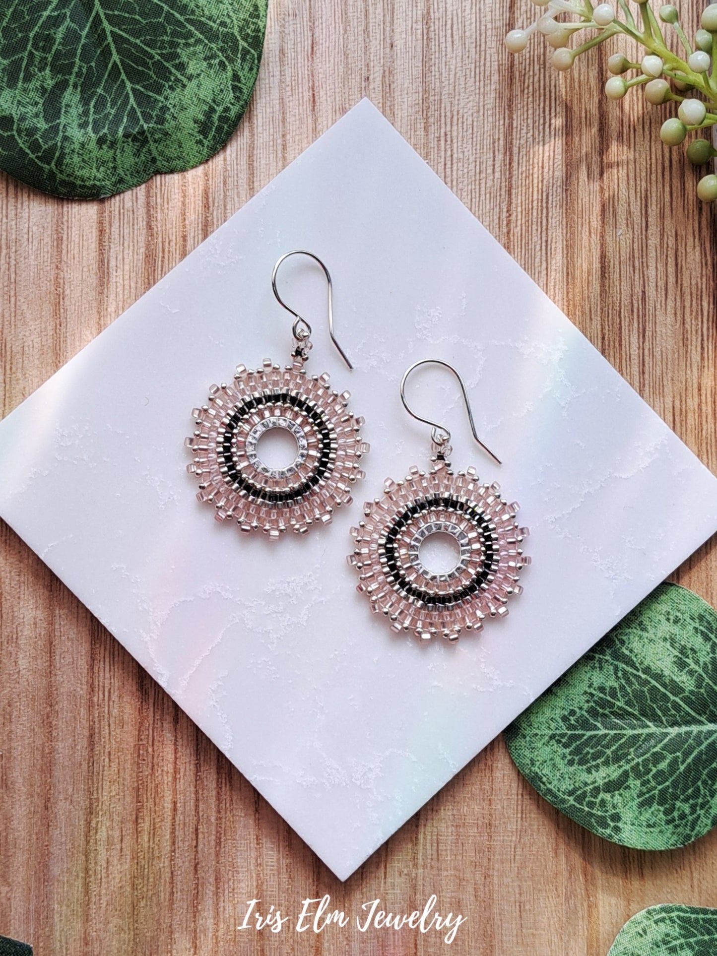Pair of pink and dark grey seed bead circle beaded earrings on a white card with green leaves in the background