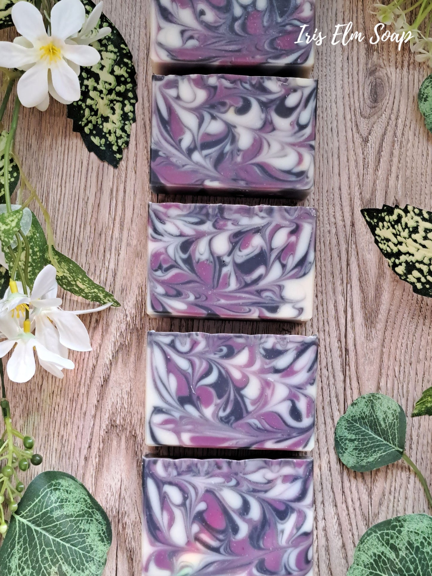 Swirl-patterned black raspberry vanilla handmade soap bars on a wooden surface with flowers and leaves.