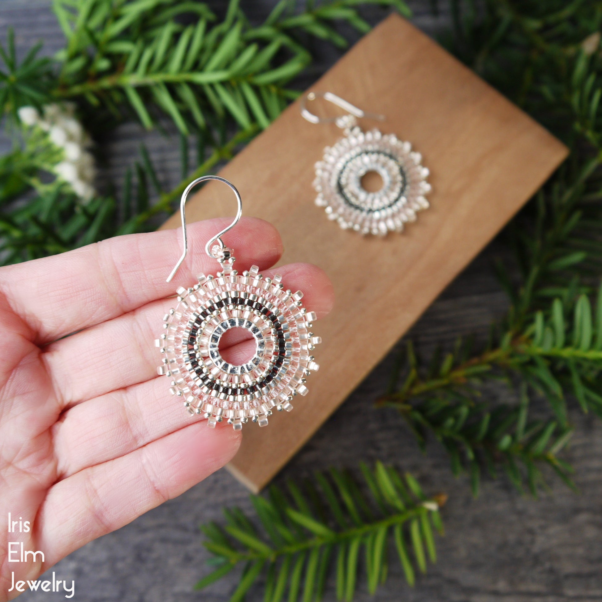 pale pink circular beaded glass earrings with sterling silver ear wires held by a hand with a wooden box and greenery in the background, featuring 'Iris Elm Jewelry' branding.