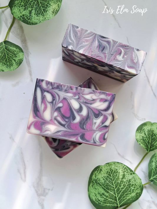 Two swirled purple and white black raspberry vanilla handmade soap bars on a white surface with green leaves.