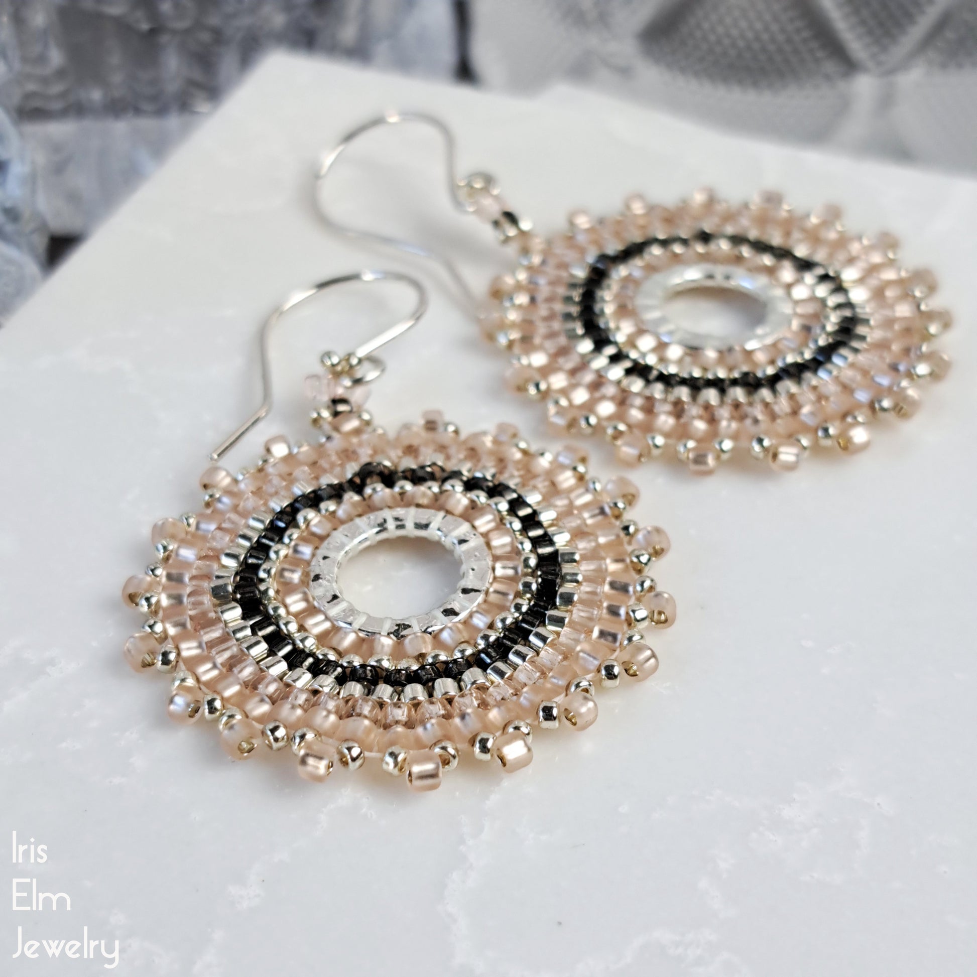 Pair of blush pink woven hoop seed bead handmade earrings on a white surface with 'Iris Elm Jewelry' branding.