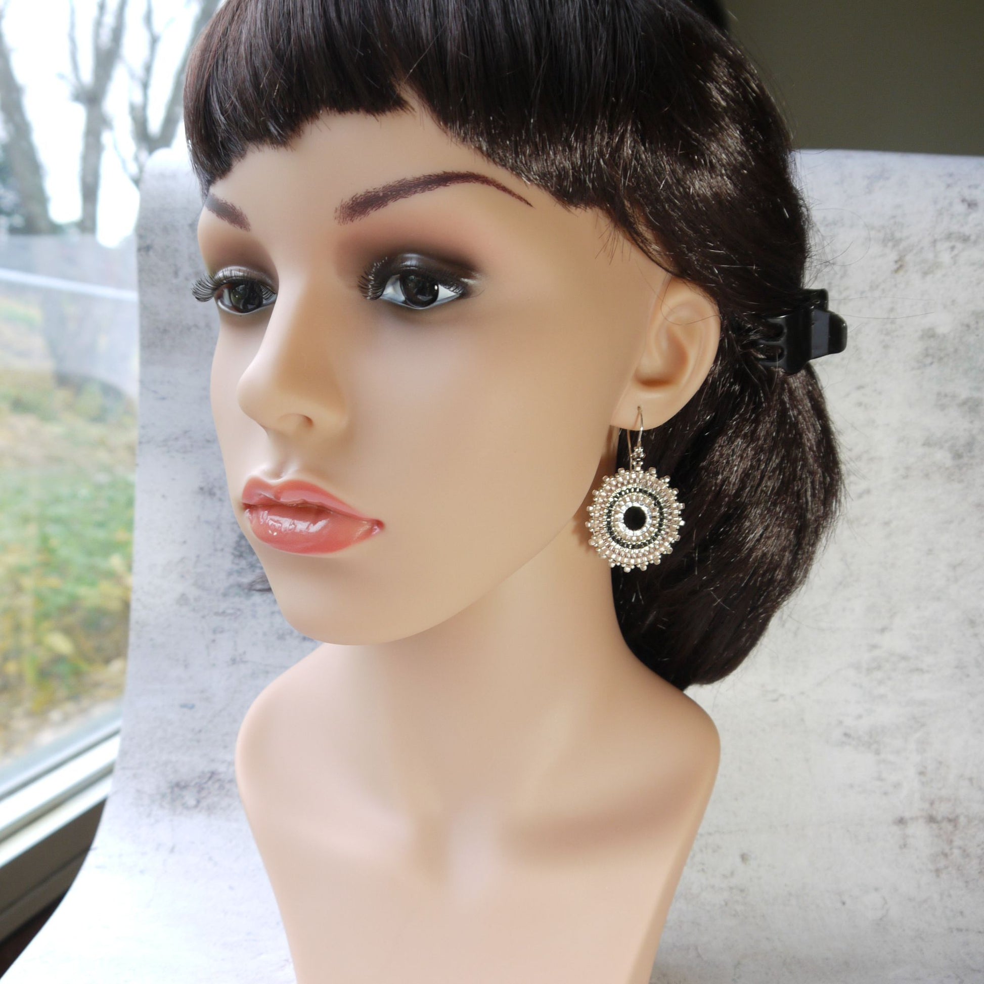 Mannequin head with dark hair and a decorative pale pink hoop seed bead earring on a blurred outdoor background