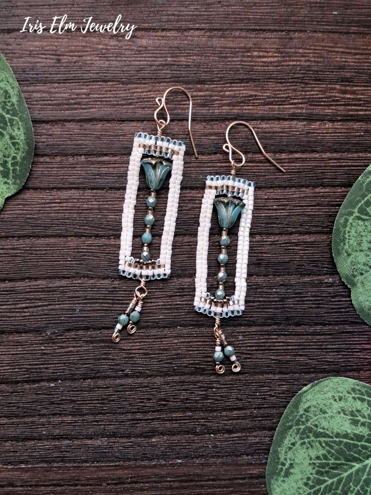 Pair of beaded earrings with tulip flower charms on a wooden surface with green leaves.