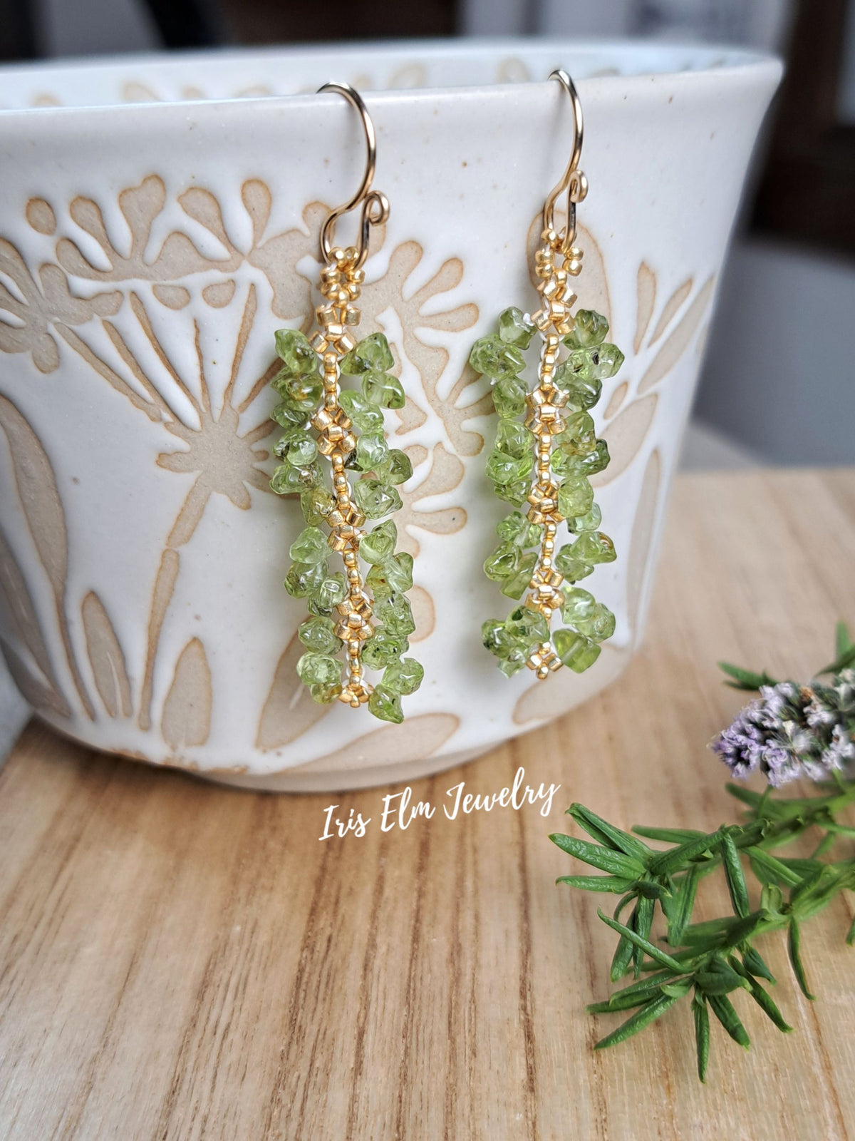 beaded green peridot chip with gold seed bead earrings by Iris Elm Jewelry dangling from ceramic pottery