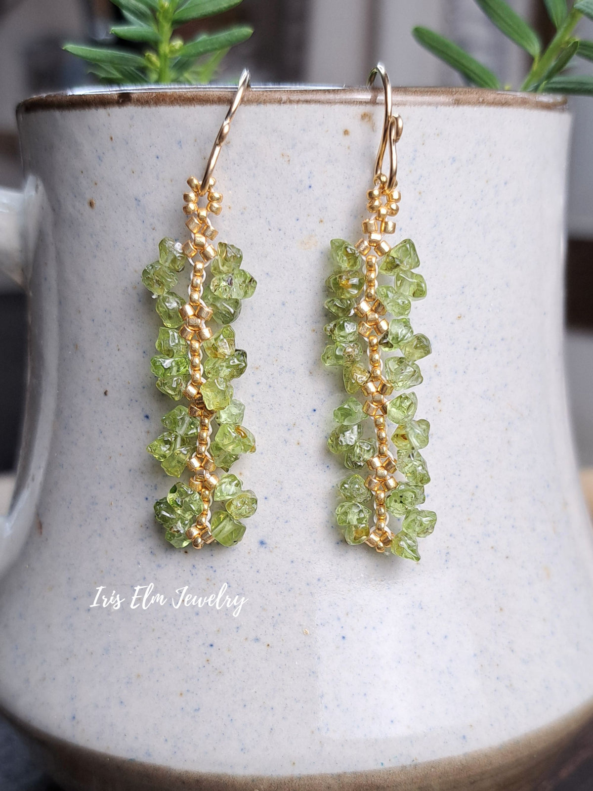 beaded green peridot chip with gold seed bead earrings by Iris Elm Jewelry dangling from ceramic pottery