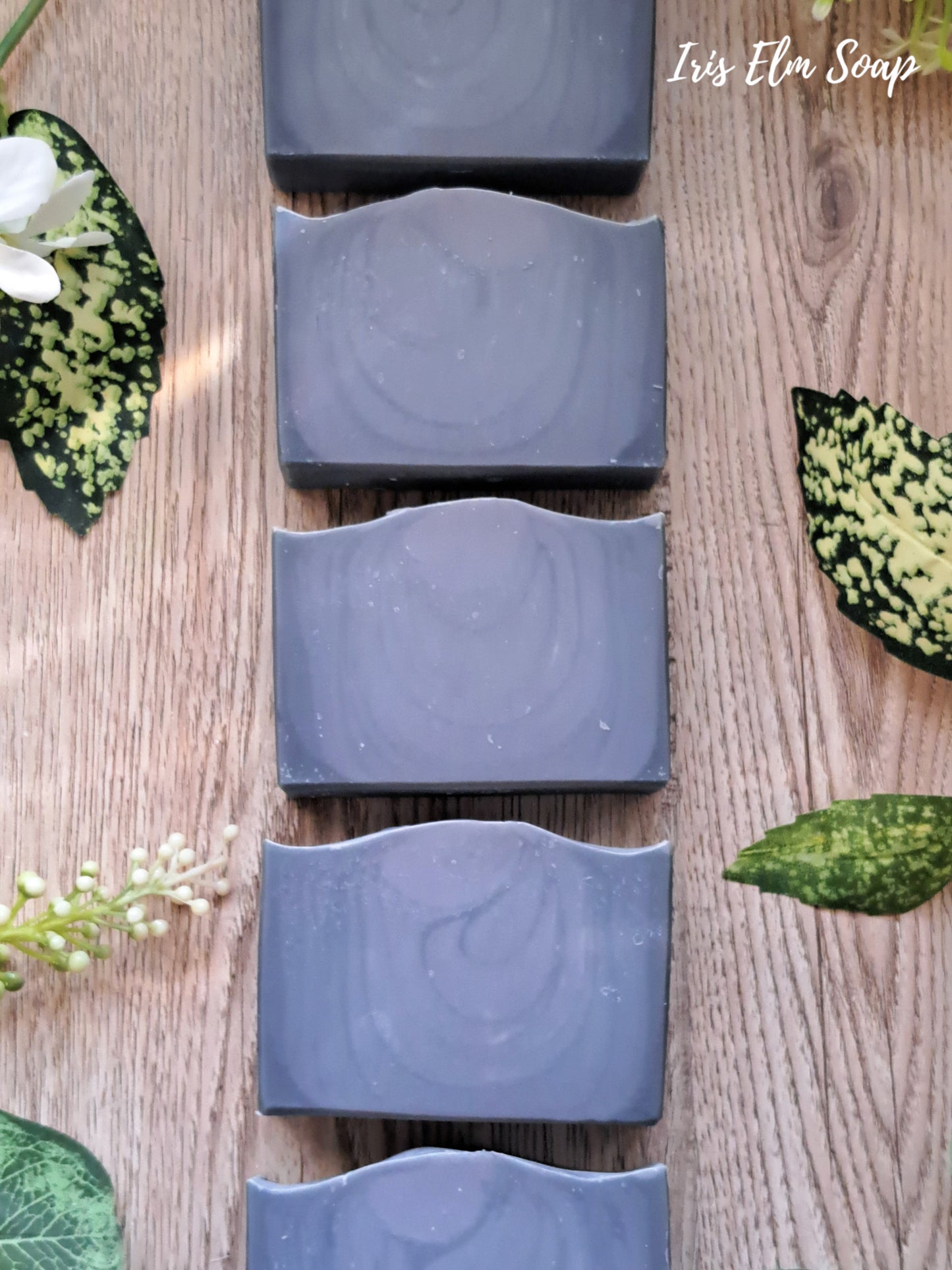 Row of dark blue cedar musk and lavender handmade soap bars on a wooden surface with green leaves and flowers.