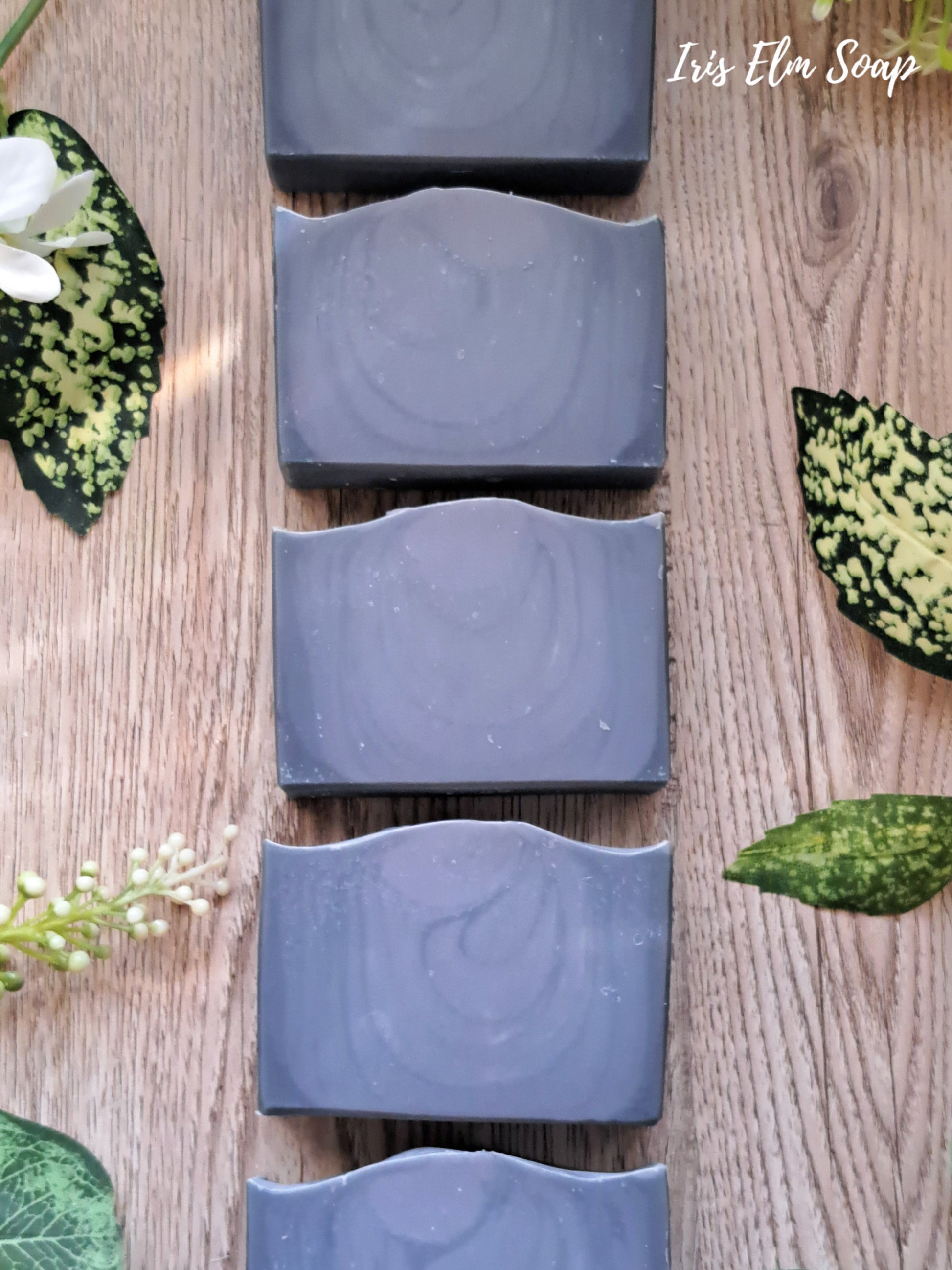 Row of dark blue cedar musk and lavender handmade soap bars on a wooden surface with green leaves and flowers.