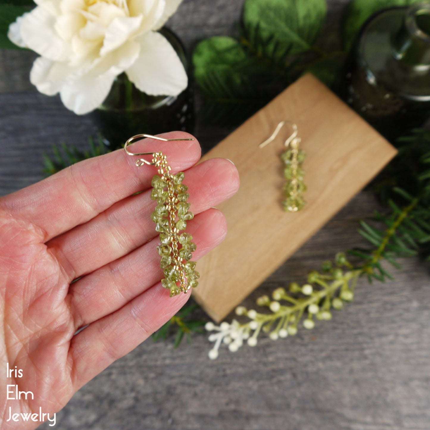 Hand holding green beaded peridot gemstone earrings with a wooden block and flowers in the background, featuring 'Iris Elm Jewelry'.