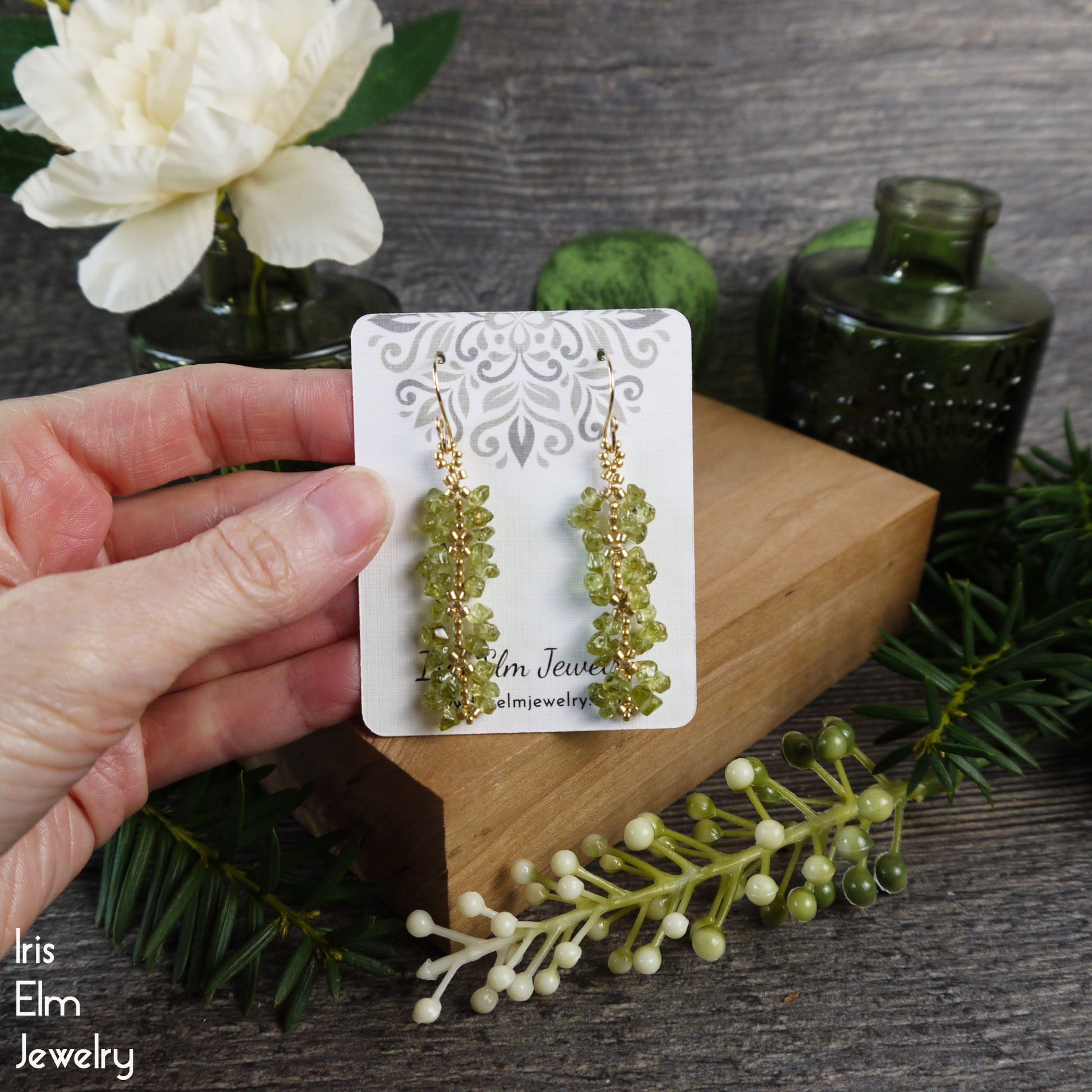 Green peridot beaded earrings on a card held by a hand with a floral and wooden background