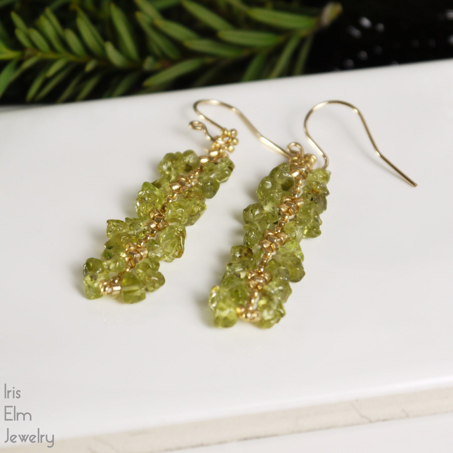 Green peridot gemstone chip beaded earrings on a white surface with a blurred green leafy background