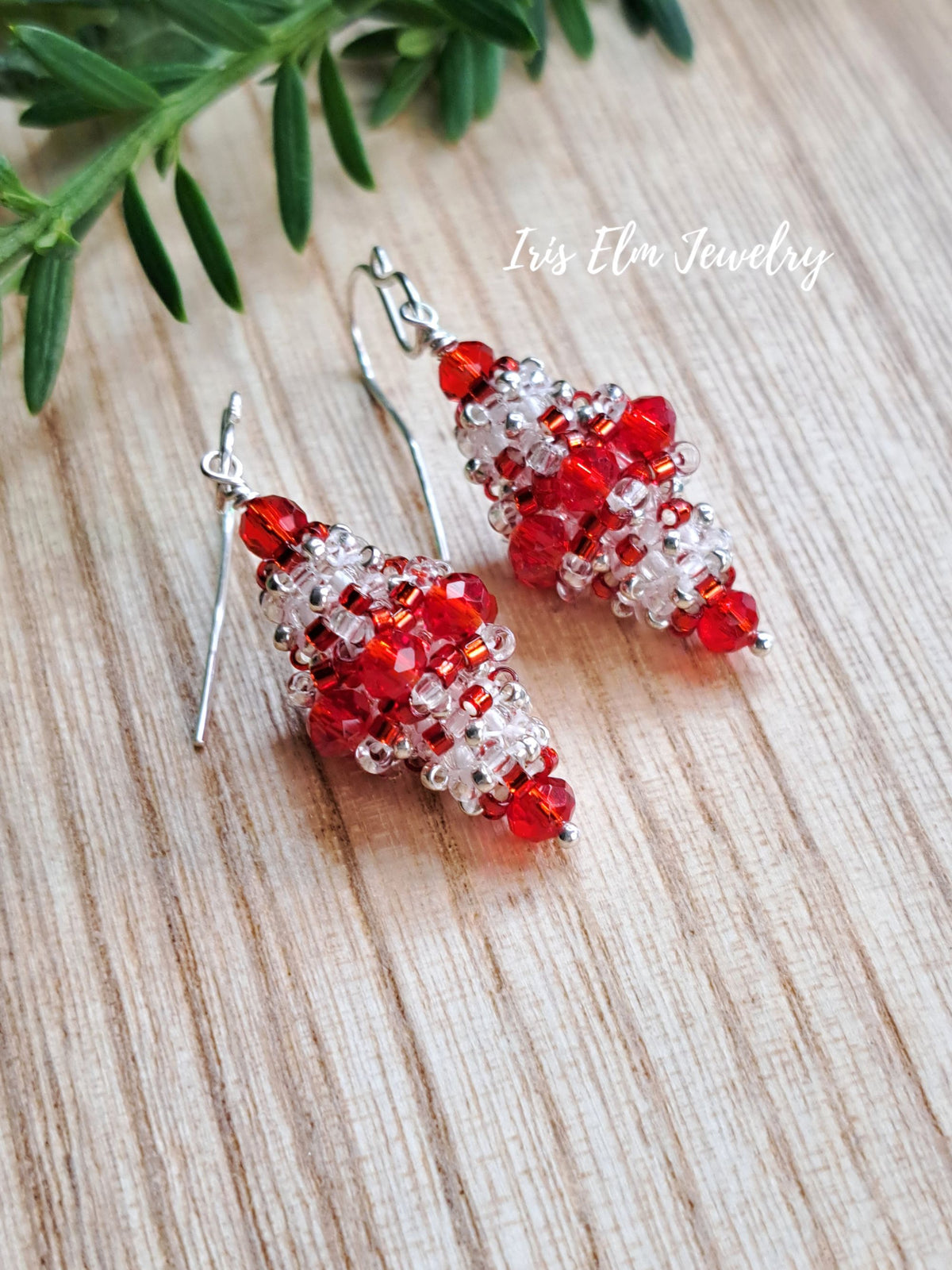 A pair of beaded dangle earrings with red and clear faceted beads on silver fishhook wires, displayed on a light wood surface with green leaves in the corner. The text 'Iris Elm Jewelry' is visible in the upper right.