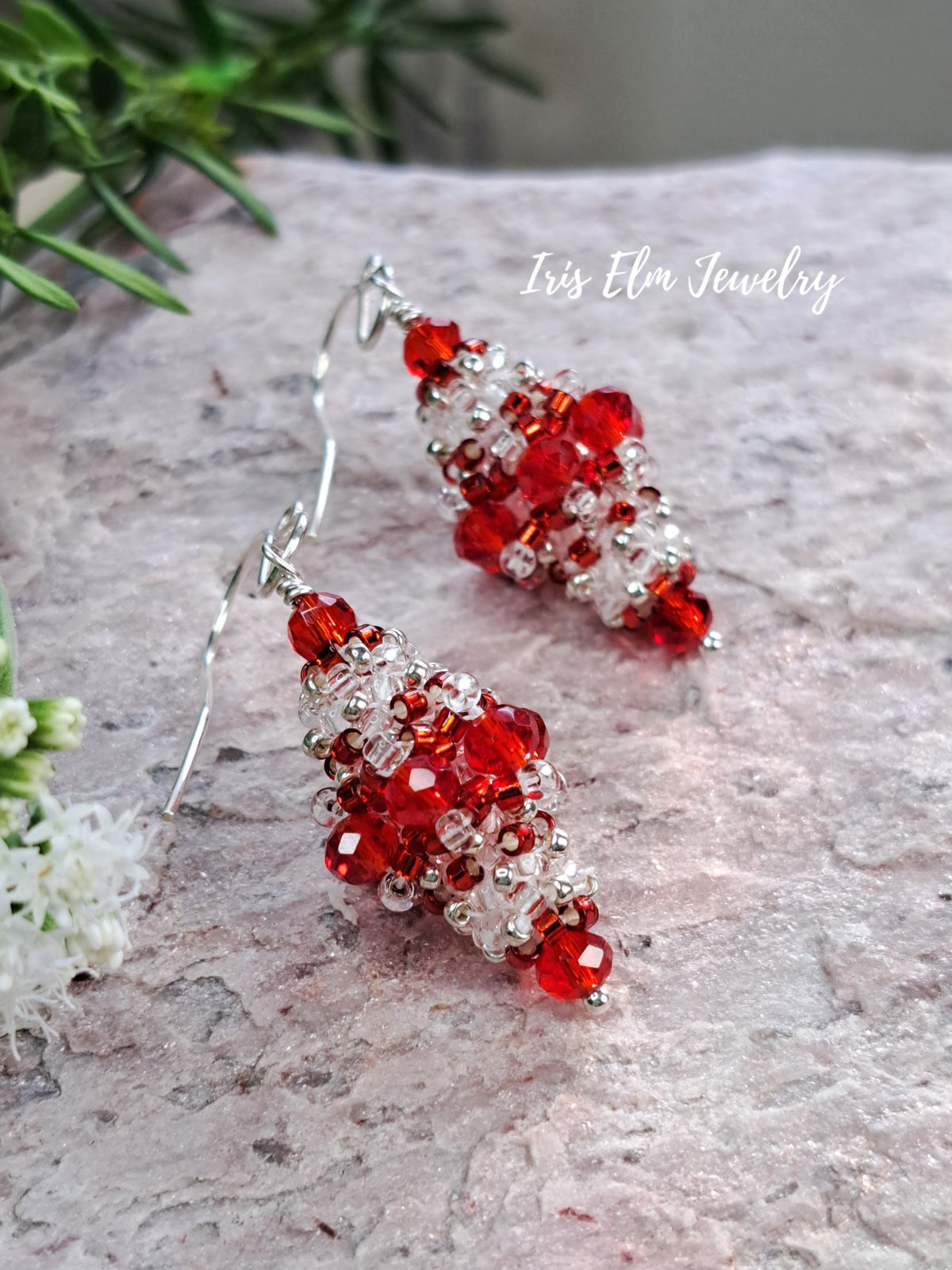 A pair of beaded dangle earrings with faceted red and clear beads on silver-toned hooks, displayed on a textured light gray stone surface with green leaves and small white flowers in the background. The text 'Iris Elm Jewelry' is visible in the upper right.