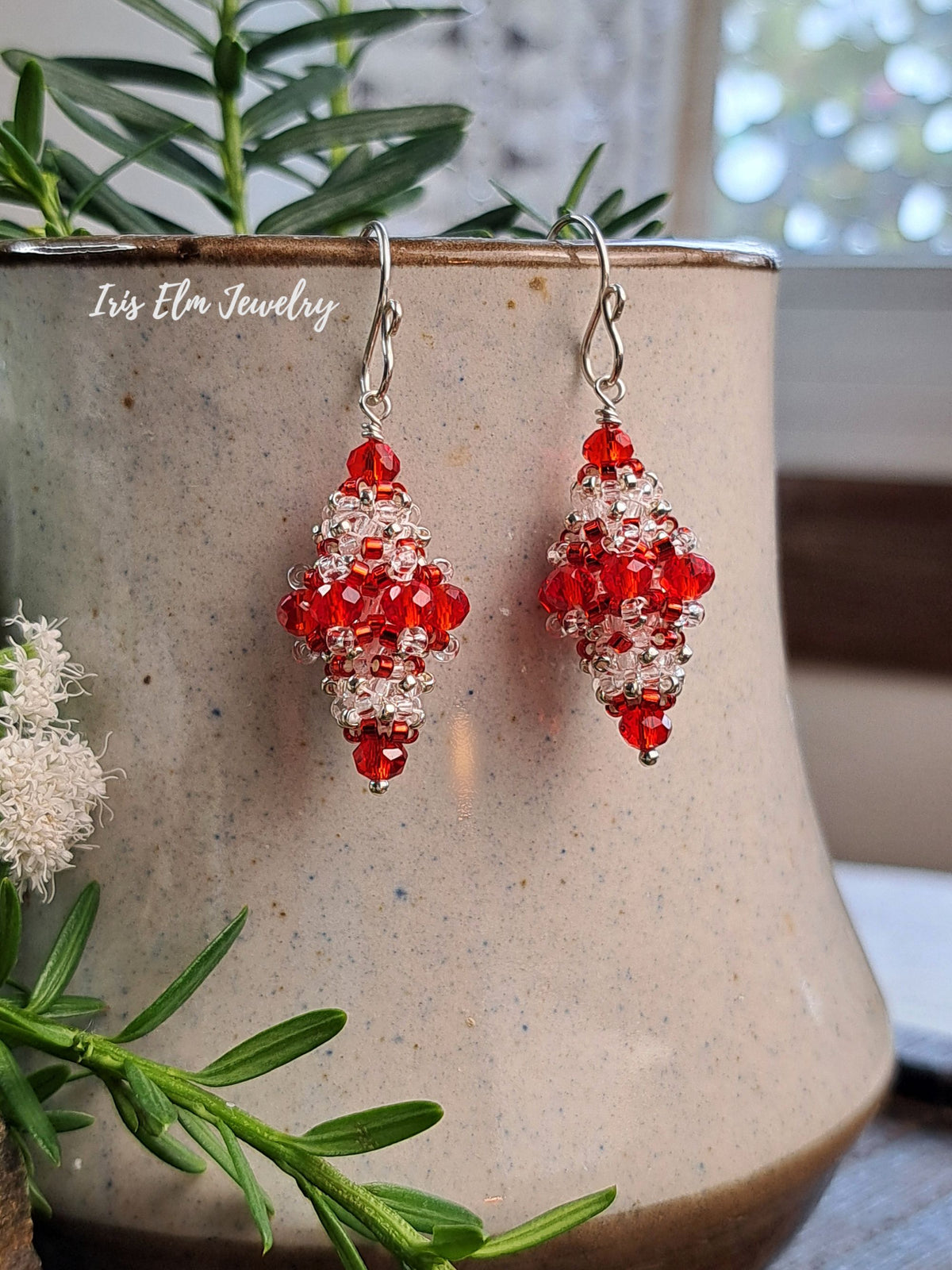 A pair of handmade beaded dangle earrings with red faceted beads and silver-toned metal hooks, displayed on a speckled beige ceramic pot with green foliage in the foreground.