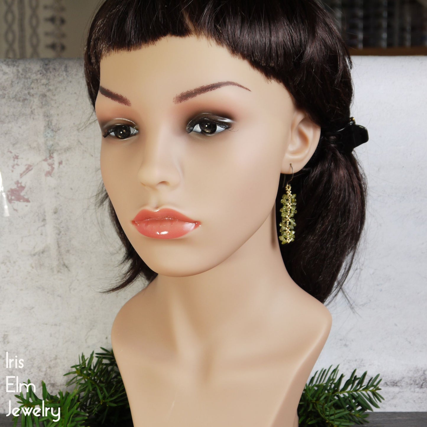 Mannequin head with dark hair and a gold green peridot earring, with 'Iris Elm Jewelry' branding.