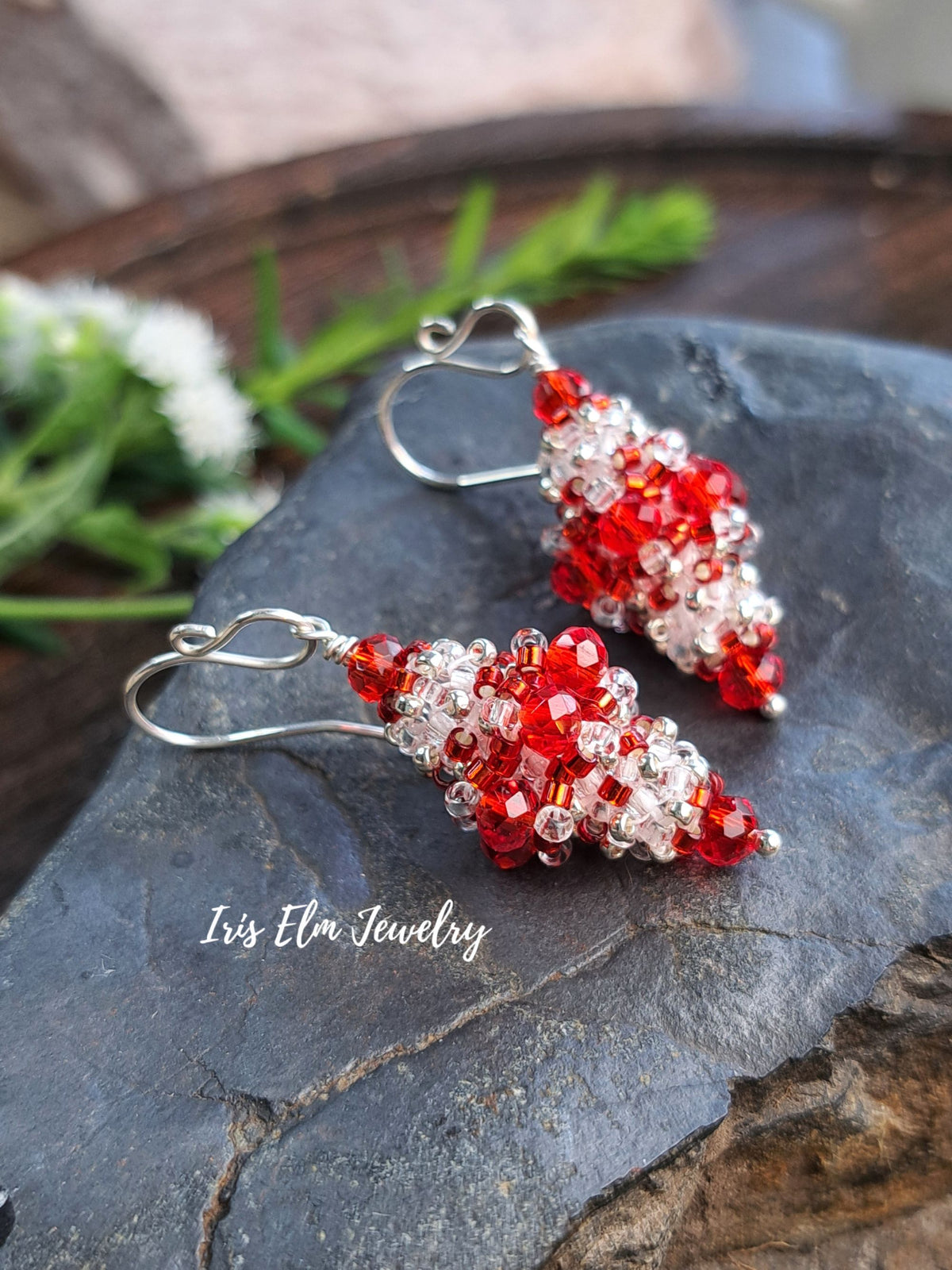 Red and clear beaded drop earrings with silver fishhook backs, displayed on a dark slate stone with green foliage in the background. The brand name 'Iris Elm Jewelry' is watermarked on the lower left.