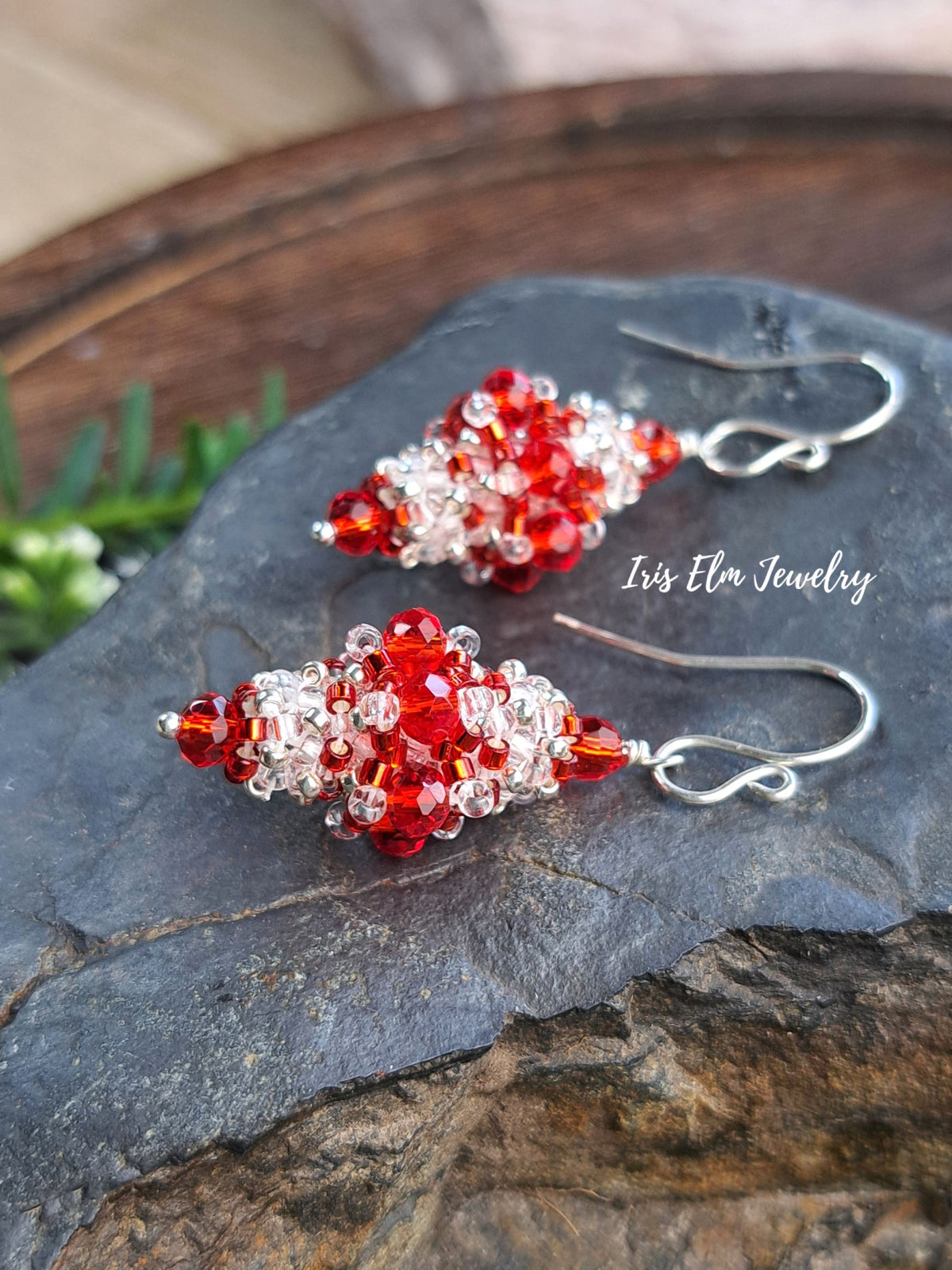 A pair of handmade beaded drop earrings with red and clear faceted beads on silver fishhook wires, displayed on a dark slate stone with a rustic wooden background. The brand name 'Iris Elm Jewelry' is watermarked on the right.