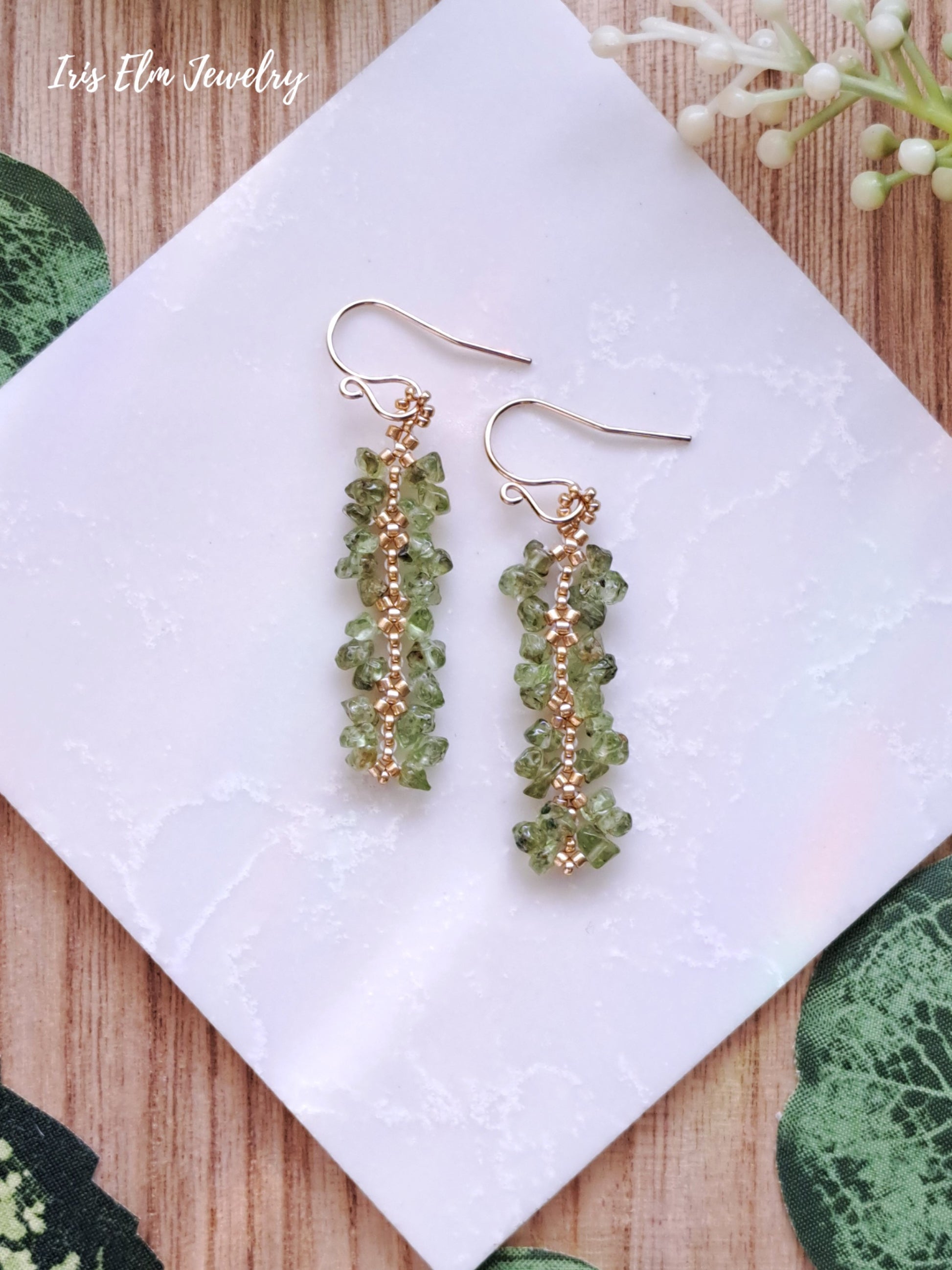 Green perdiot and gold seed bead beaded earrings on a white napkin with 'Iris Elm Jewelry' branding.