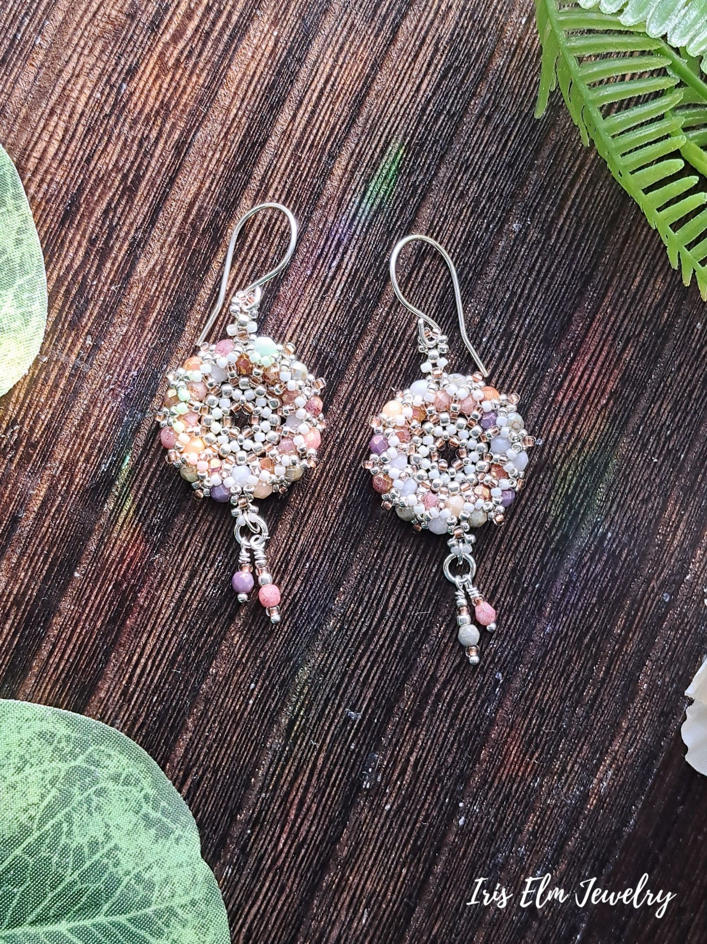 Pair of beaded multi color czech glass woven wreath hoop earrings on a wooden surface with green leaves.