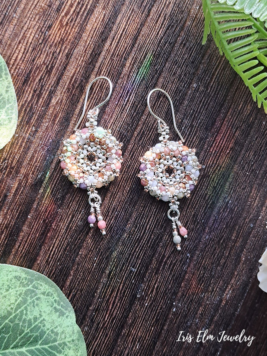 Pair of beaded multi color czech glass woven wreath hoop earrings on a wooden surface with green leaves.