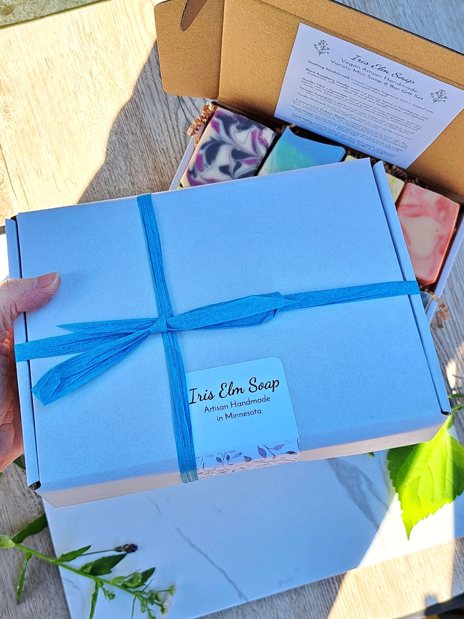 Blue gift box with a blue ribbon held by a hand, with 'Iris Elm Soap' branding.
