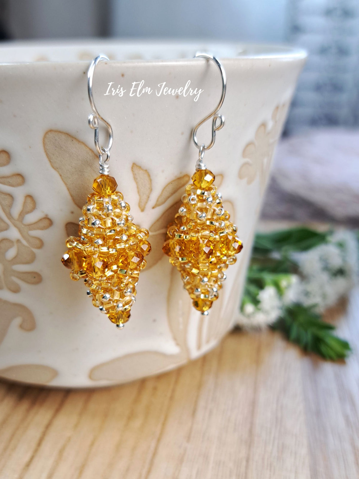 A pair of handmade beaded dangle earrings in amber yellow and silver, hanging from a ceramic mug on a wooden table with a blurred background.
