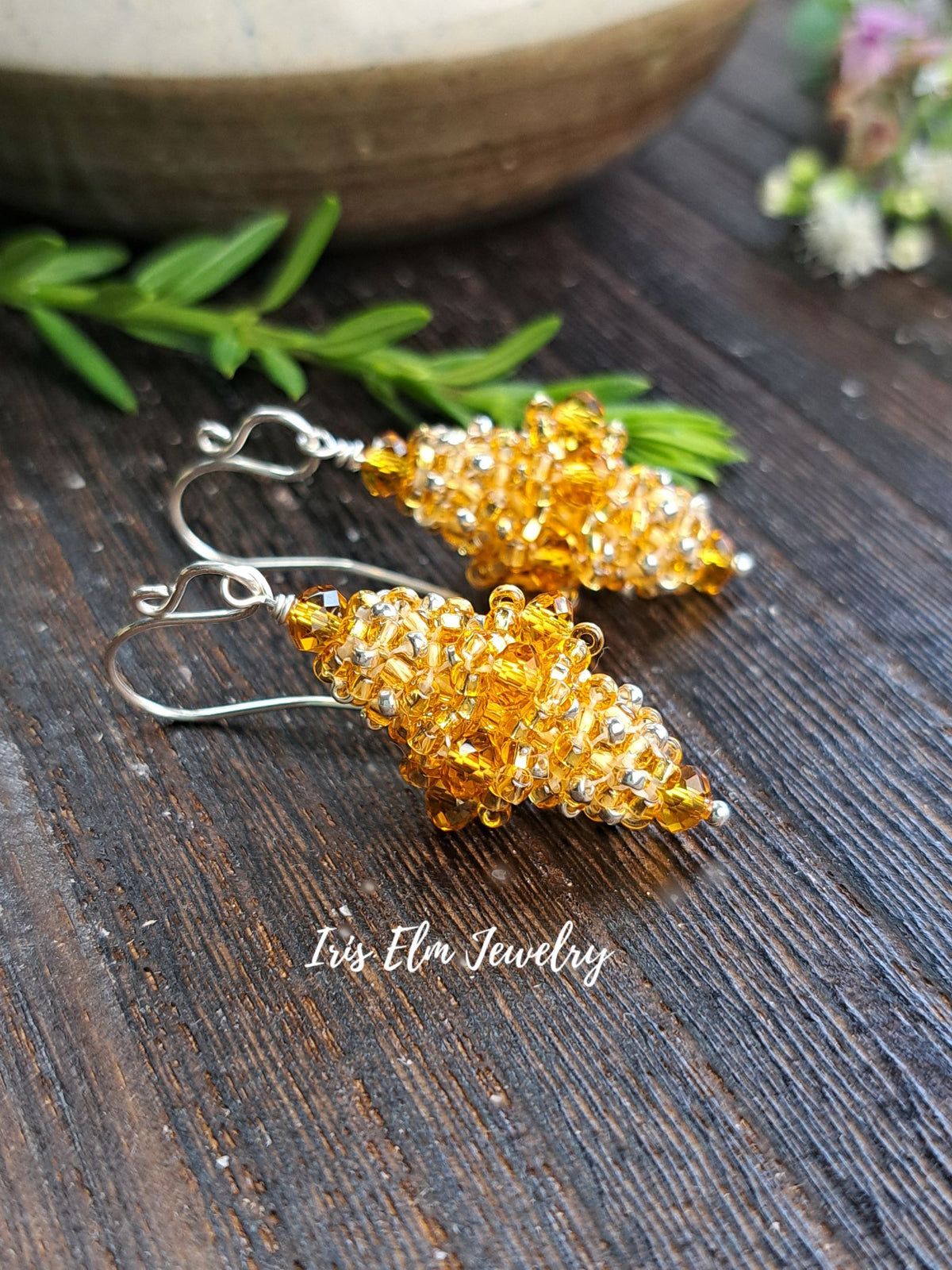 A pair of handmade amber beaded dangle earrings with silver fishhook wires, displayed on a dark rustic wooden surface with green rosemary sprigs and a ceramic bowl in the background. The brand name 'Iris Elm Jewelry' is watermarked at the bottom.