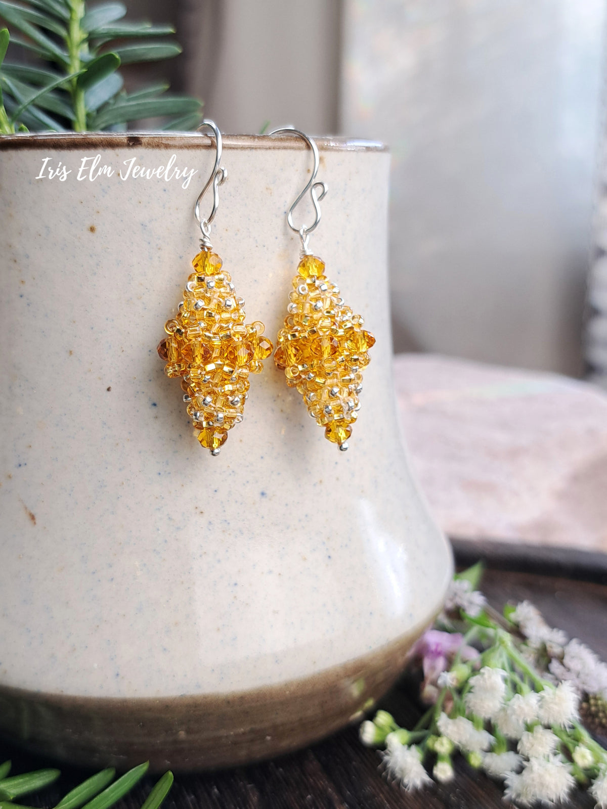 A pair of handmade dangle earrings by Iris Elm Jewelry, featuring textured amber-yellow crystal clusters on silver French hook wires, displayed on a rustic ceramic pot.