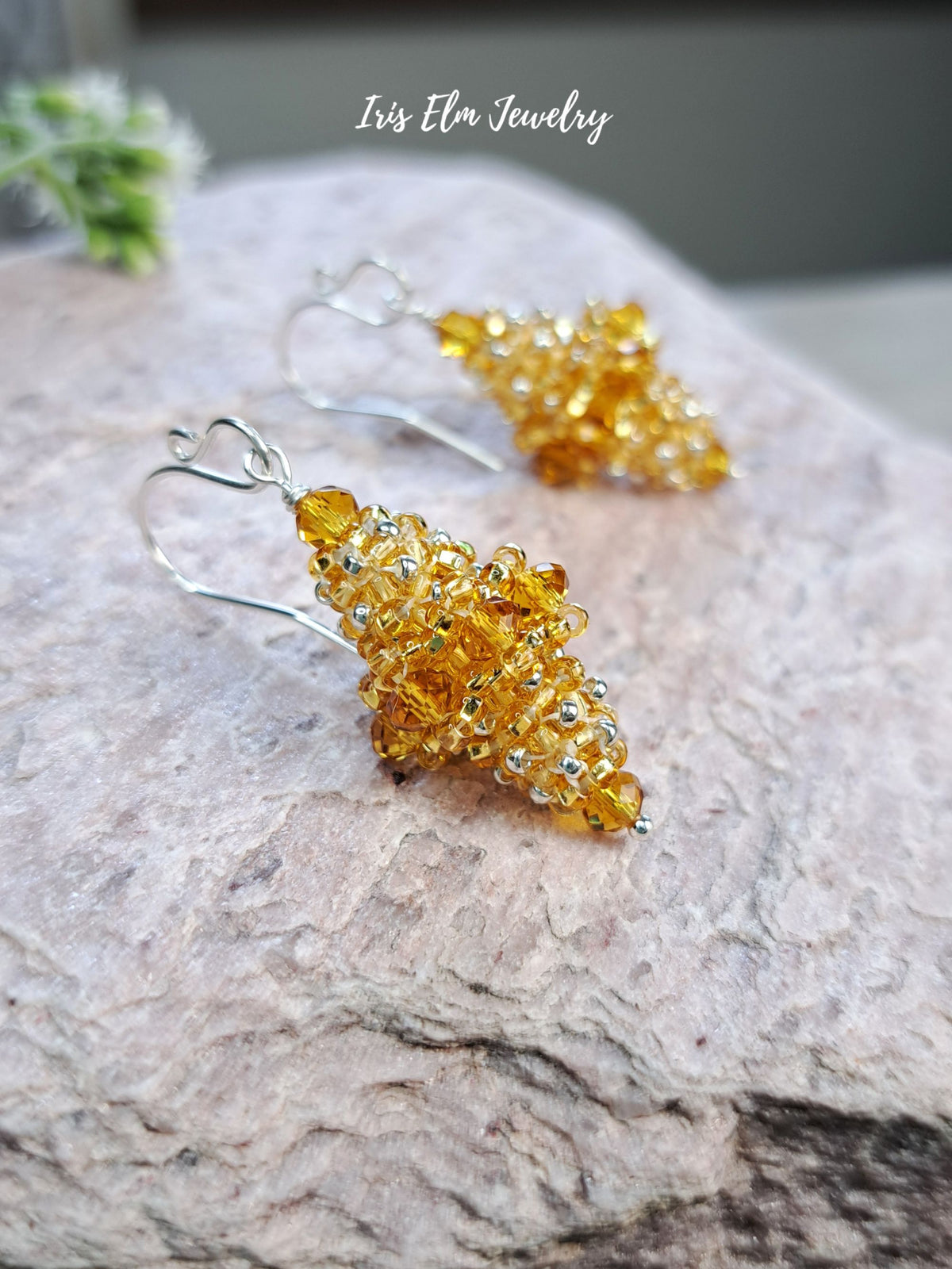A pair of amber-colored beaded dangle earrings with silver fishhook wires, displayed on a textured light gray stone surface. The brand name 'Iris Elm Jewelry' is written in white script at the top.