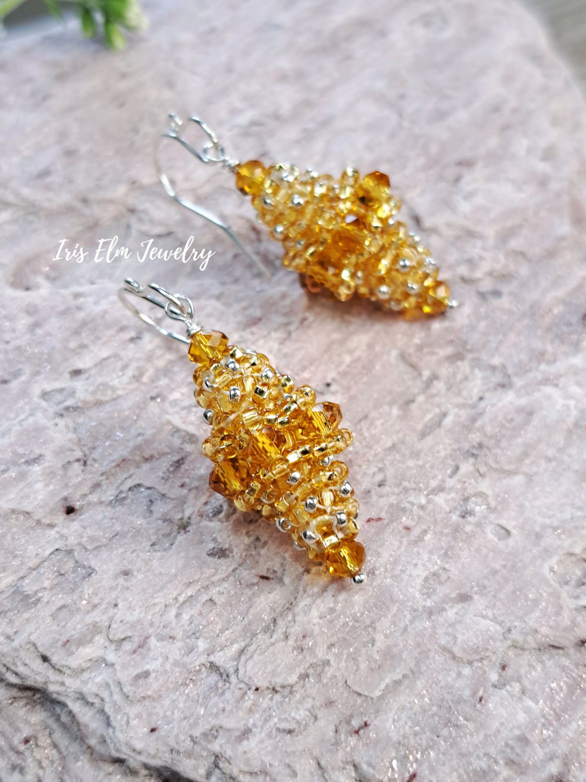 A pair of handmade amber and crystal cluster dangle earrings with silver hooks, displayed on a textured light gray stone surface. A watermark for 'Iris Elm Jewelry' is visible on the left.