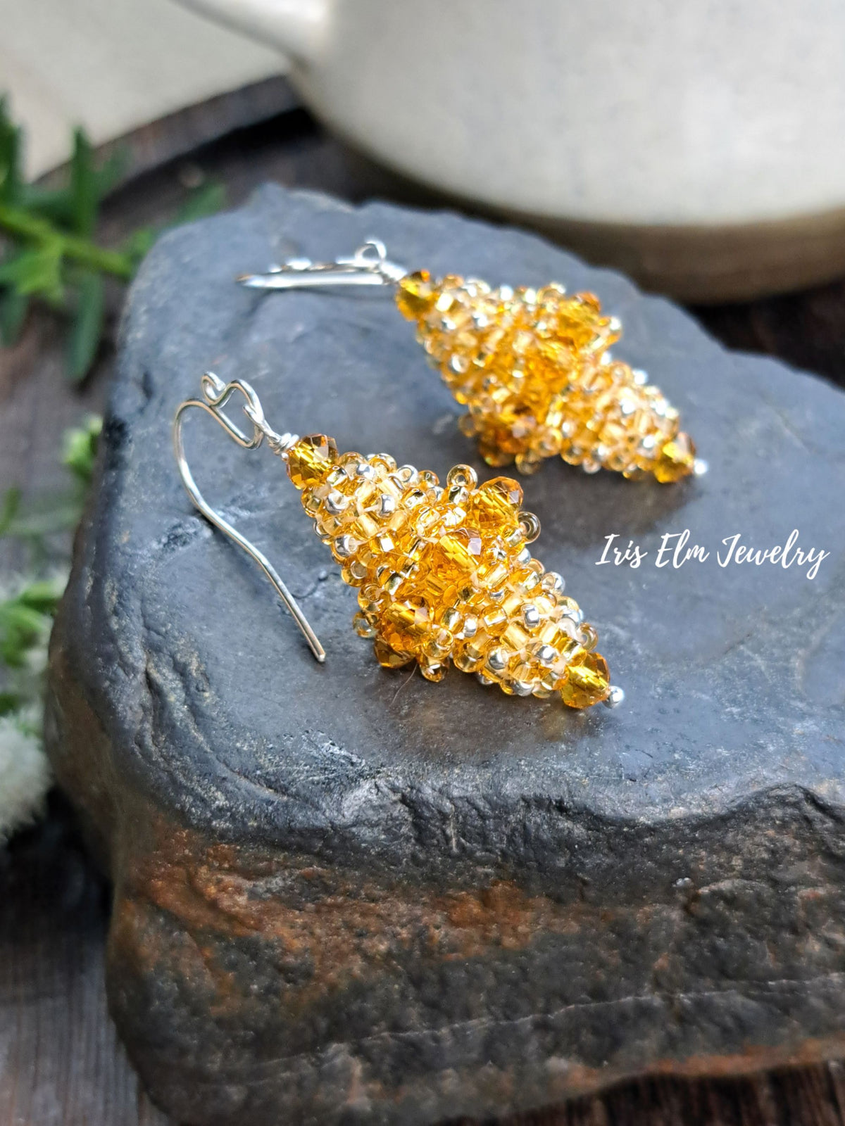 A pair of topaz yellow glass and silver beaded dangle earrings displayed on a dark textured stone surface, with a ceramic bowl and green leaves in the background. The brand name 'Iris Elm Jewelry' is watermarked on the right.