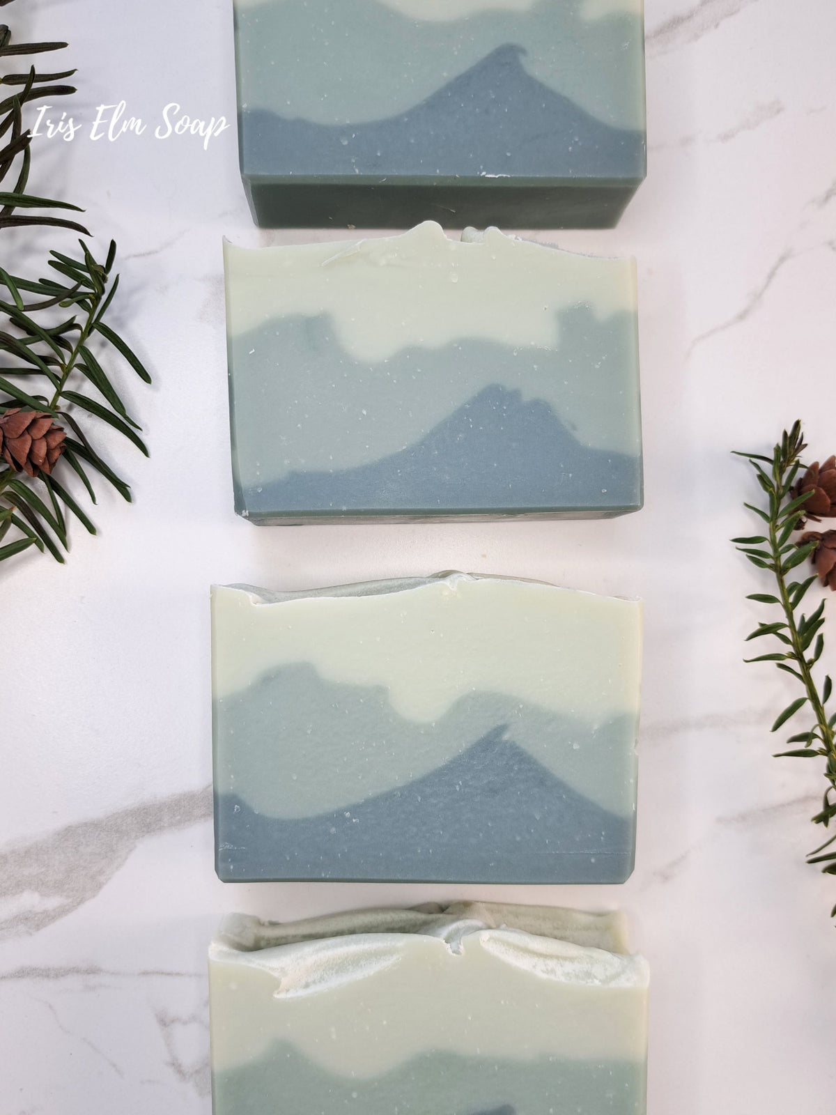 Four rectangular bars of layered balsam fir soap with a mountain wave pattern in shades of slate blue and cream, arranged vertically on a white marble surface with pine branches and pinecones as decorative props.