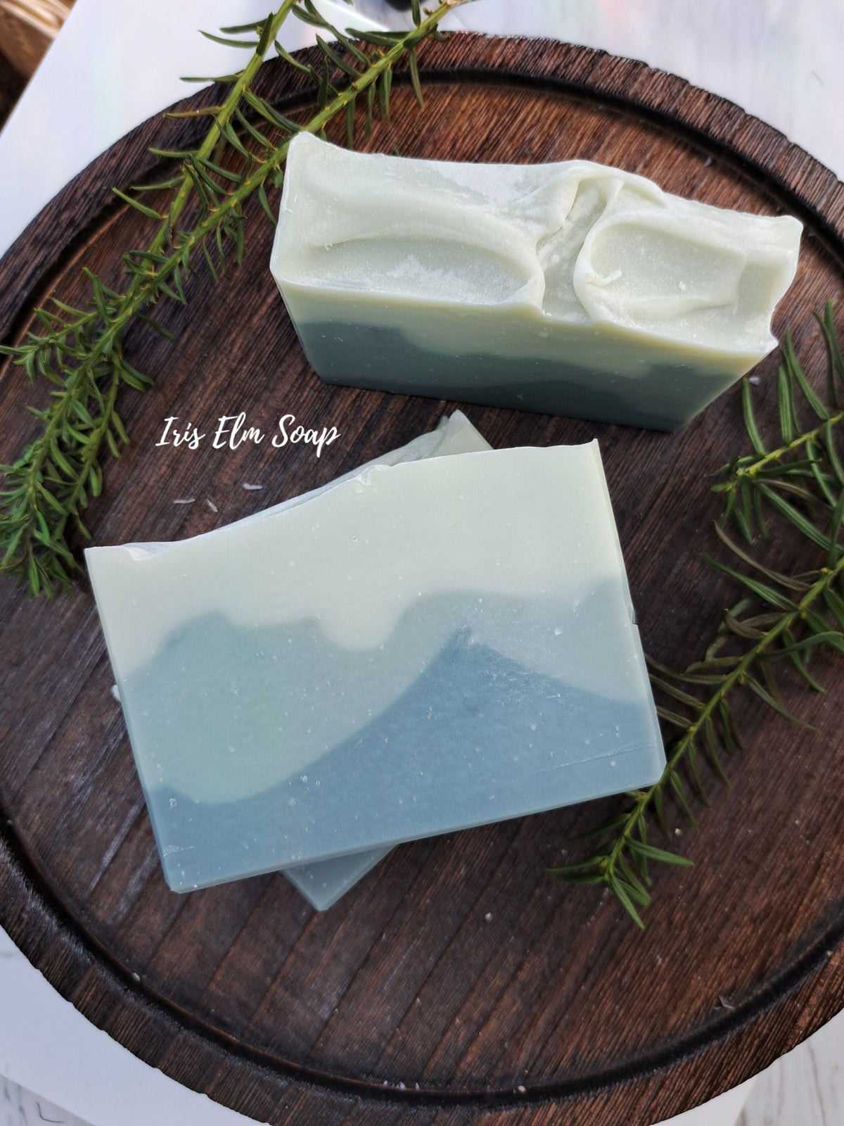 Two handmade layered balsam fir soap bars with a sage green and light blue marbled design, displayed on a dark wooden tray with rosemary sprigs. The brand name 'Iris Elm Soap' is printed on the wooden tray.