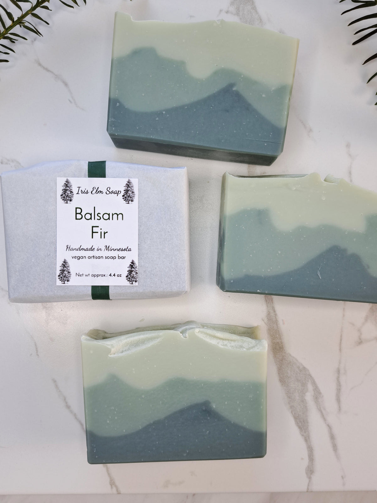 Four bars of artisan soap with a mountain-like layered design in sage green and slate blue, displayed on a white marble surface alongside a wrapped bar labeled 'Balsam Fir' by Iris Elm Soap.