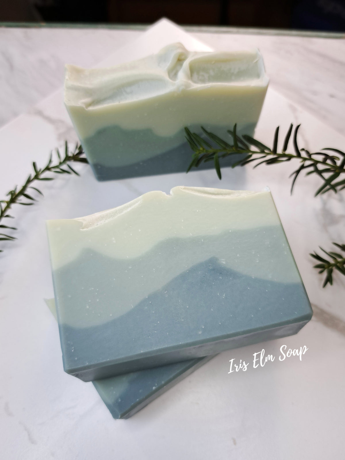 Two balsam fir handmade layered soap bars with a wavy mountain pattern in pale green and light blue, styled on a white marble surface with rosemary sprigs and a brand watermark 'Iris Elm Soap'.
