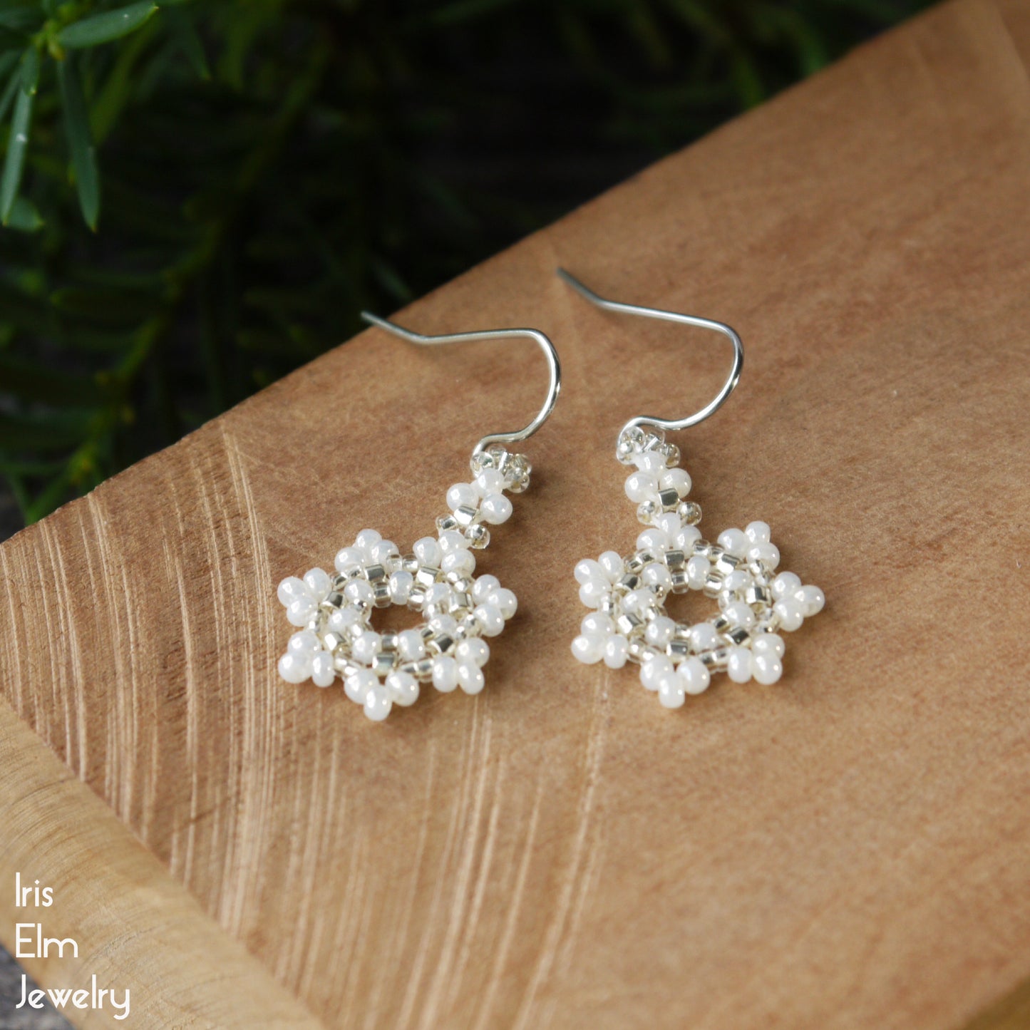 Pair of small seed bead snowflake earrings with pearl-like beads on a wooden surface, branded 'Iris Elm Jewelry'.