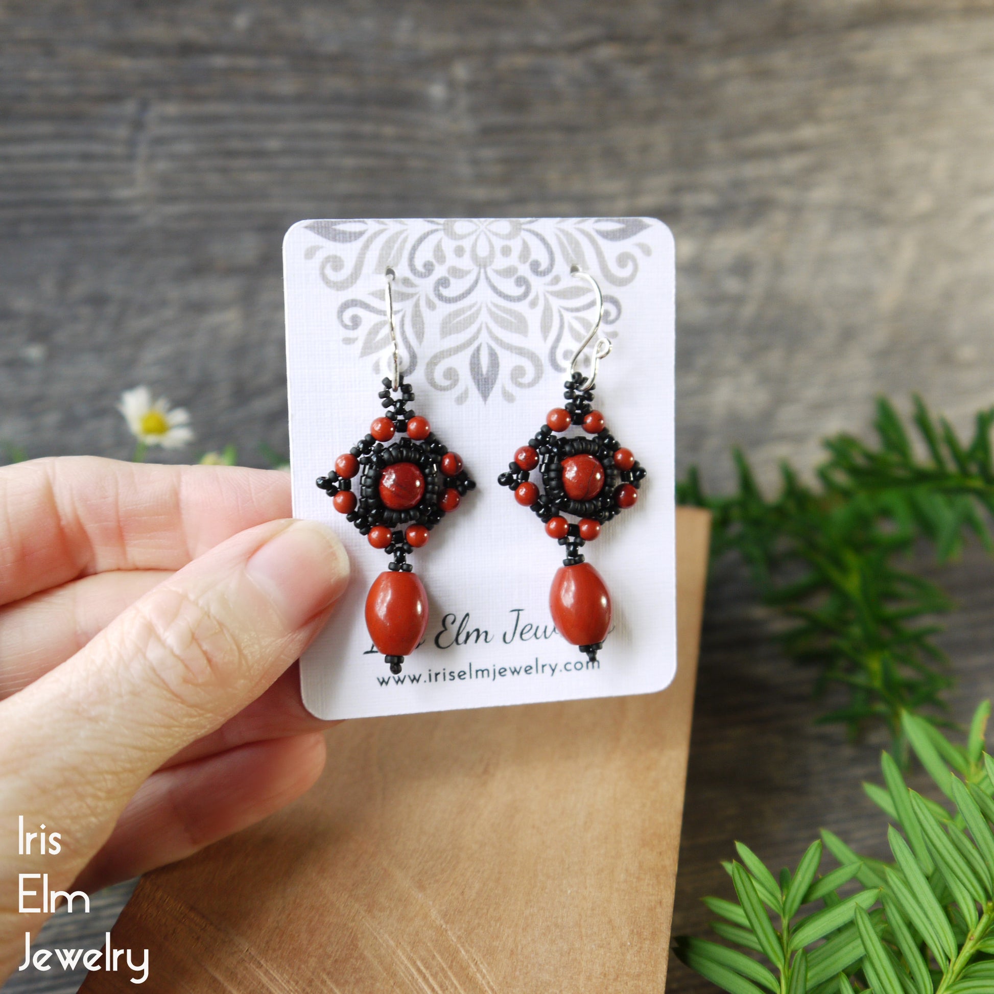 handmade red jasper earrings with black beaded accents on a 'Iris Elm Jewelry' card held by a hand with a wooden background.