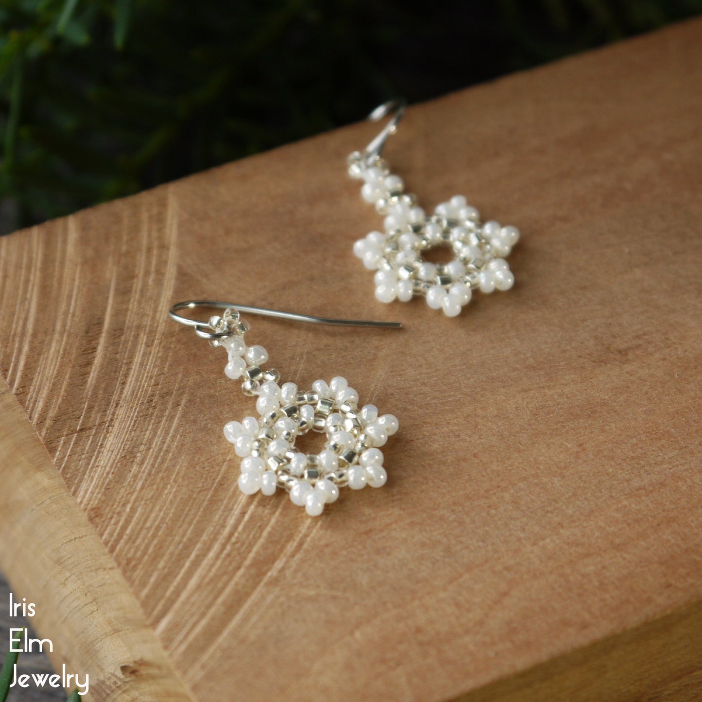 Pearl white seed bead snowflake handmade earrings on a wooden surface with 'Iris Elm Jewelry' branding.