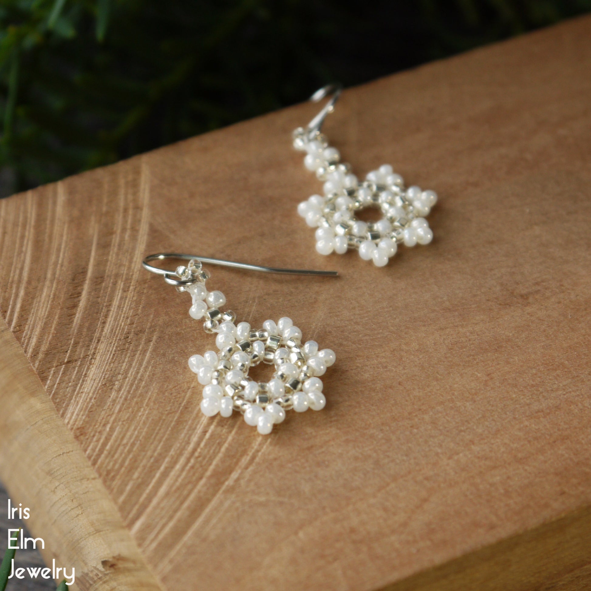Pearl white seed bead snowflake handmade earrings on a wooden surface with 'Iris Elm Jewelry' branding.