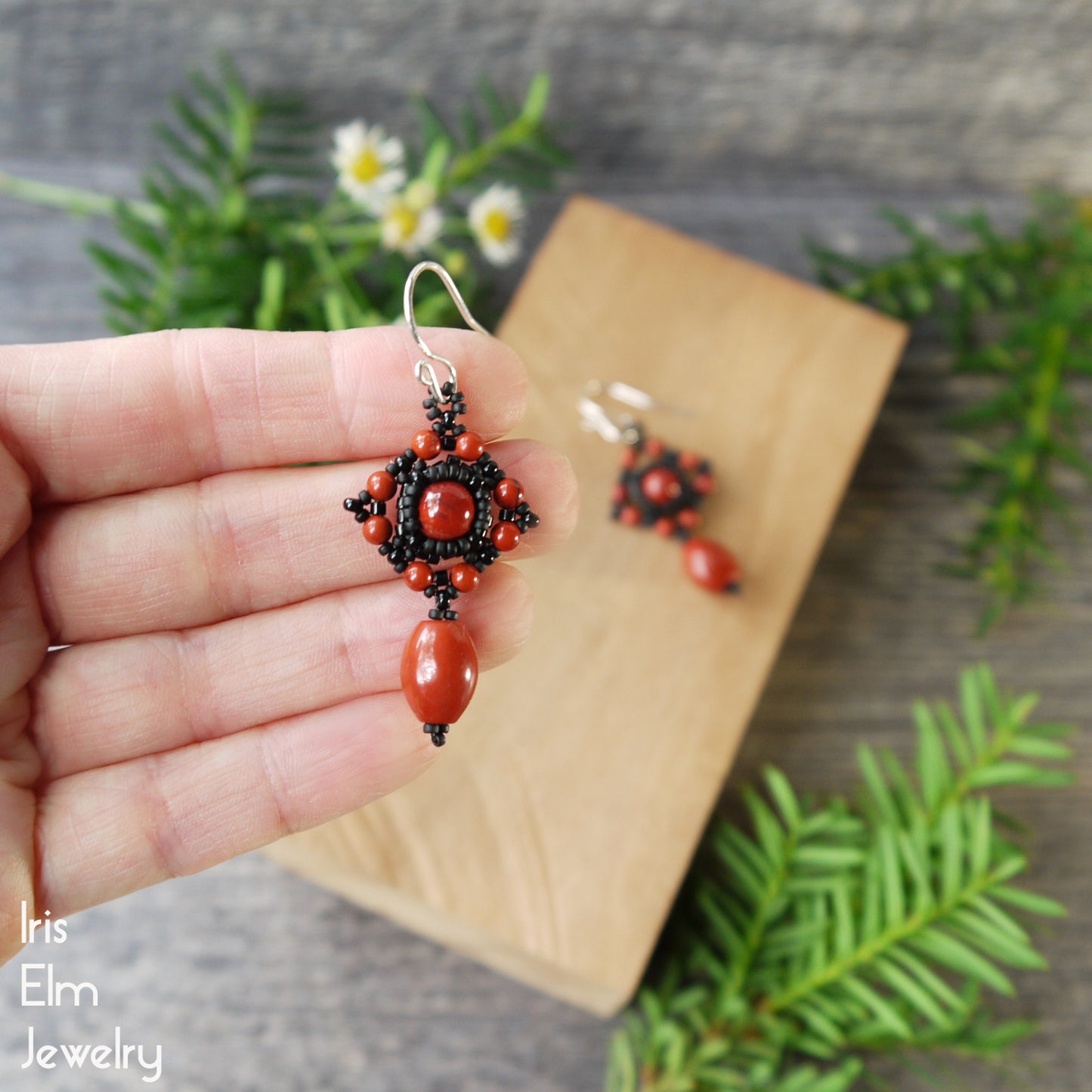 Hand holding a pair of red jasper and black beaded earrings with a natural background